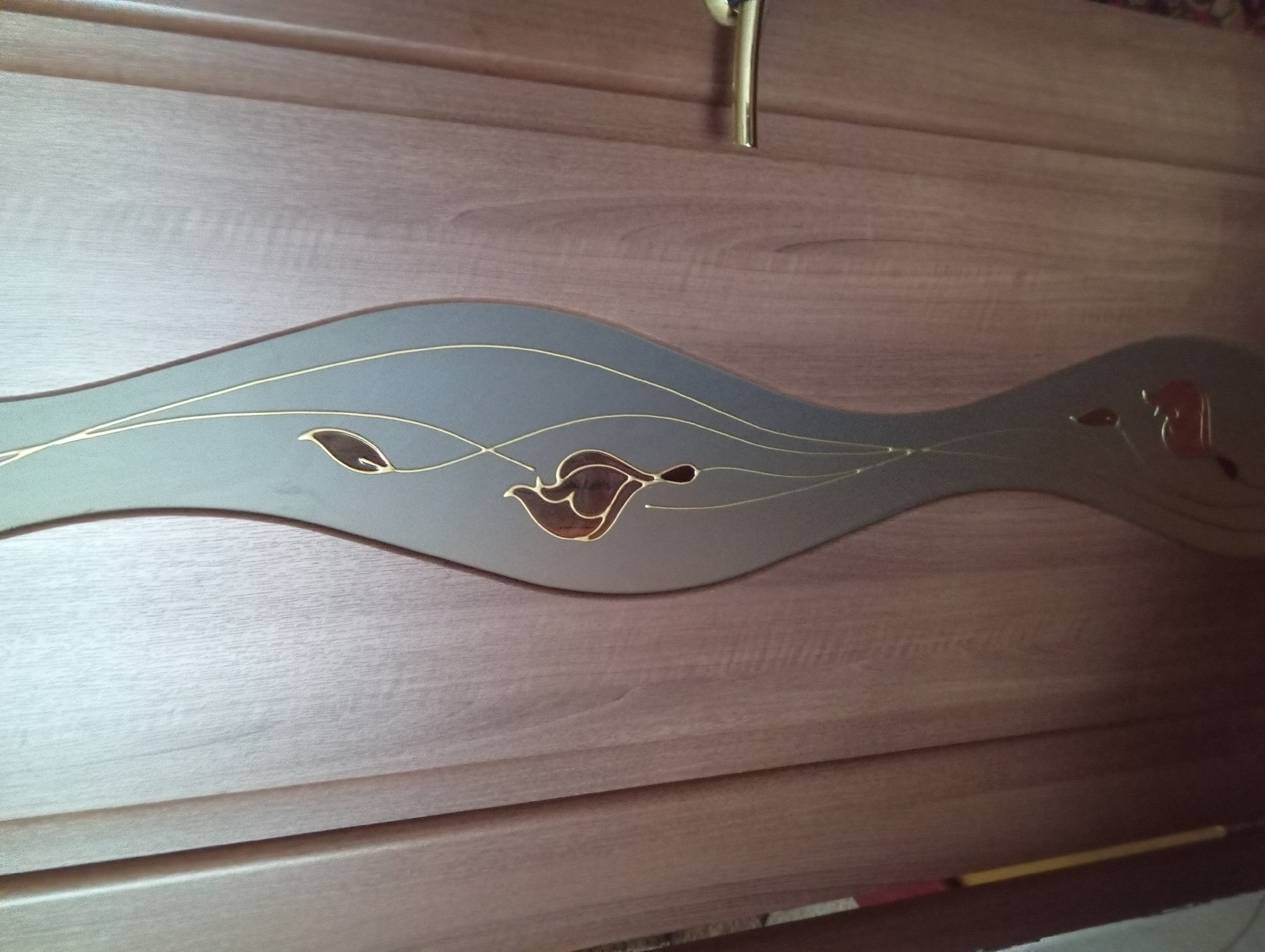 The image shows a close-up of a wooden door with a decorative inlay. The inlay is a wavy, vertical panel with a gray background and gold-colored lines forming abstract floral or leaf shapes. One of the shapes is a dark brown, leaf-like figure outlined in gold. The door appears to be a light brown wood grain. A gold-colored door handle is visible on the right side of the door. The lighting suggests an indoor environment, likely a home or office. The specific location in Kremenchuk, Ukraine, is not discernible from the image itself.