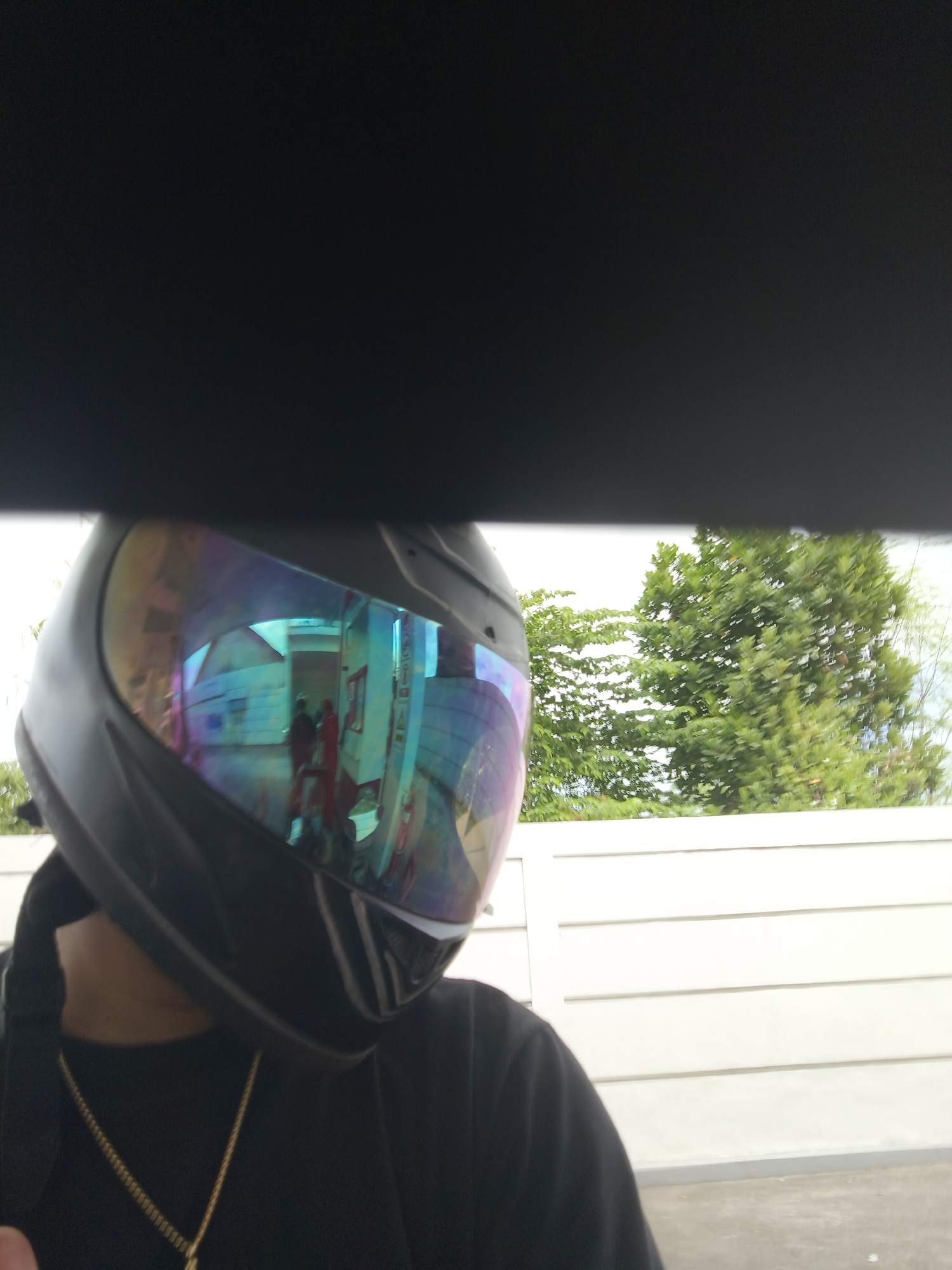 The image shows a person wearing a black motorcycle helmet with a highly reflective visor. The visor reflects an indoor scene with white tiled walls and several people, some wearing red clothing. Outside the helmet, a white paneled wall and green trees are visible against a bright sky, suggesting an outdoor setting. The context suggests this photo was taken in Denpasar, Indonesia, likely near a building with this type of outdoor wall and lush vegetation, characteristic of the city's residential or commercial areas. The person is wearing a black shirt and a gold chain. The overall impression is a candid shot, possibly taken by the individual themselves, showcasing the reflections in their helmet.