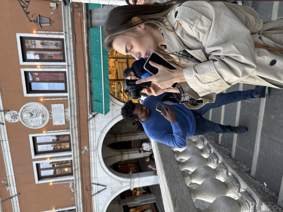 The image depicts a street scene on the Rialto Bridge in Venice, Italy. In the immediate foreground, a light-skinned female with long, wavy brown hair, dressed in a light tan trench coat over a plaid shirt, stands on stone steps behind a gray stone balustrade. She holds a black digital camera with her right hand, raising it to eye level, while simultaneously holding a silver-cased smartphone in her left hand, positioned below the camera. Her gaze is directed forward, and her lips are slightly pursed.
Behind her, on the same stone steps, a dark-skinned male with short, curly black hair, wearing a bright blue crew-neck sweater and dark blue jeans, descends the steps. He holds a silver smartphone in his left hand, looking at its screen with a slight smile.
The background features a prominent reddish-orange building with white architectural details. On its upper floor, there are three dark-framed rectangular windows. Between the two left windows, a large, circular white stone relief carving of the winged Lion of Saint Mark holding a book is affixed. Below this relief, a rectangular stone plaque with Latin inscriptions, including the partially visible date "MDXXIV" (1524), is present. Two vertical orange decorative lights flank the relief and plaque. The ground floor of the background building consists of multiple arched openings, revealing brightly lit shops with merchandise and several indistinct background figures. A green awning extends over one of these shops. A black, ornate street lamp is visible on the far right of the background structure. The scene conveys a public space with individuals engaged in photography and personal device use.