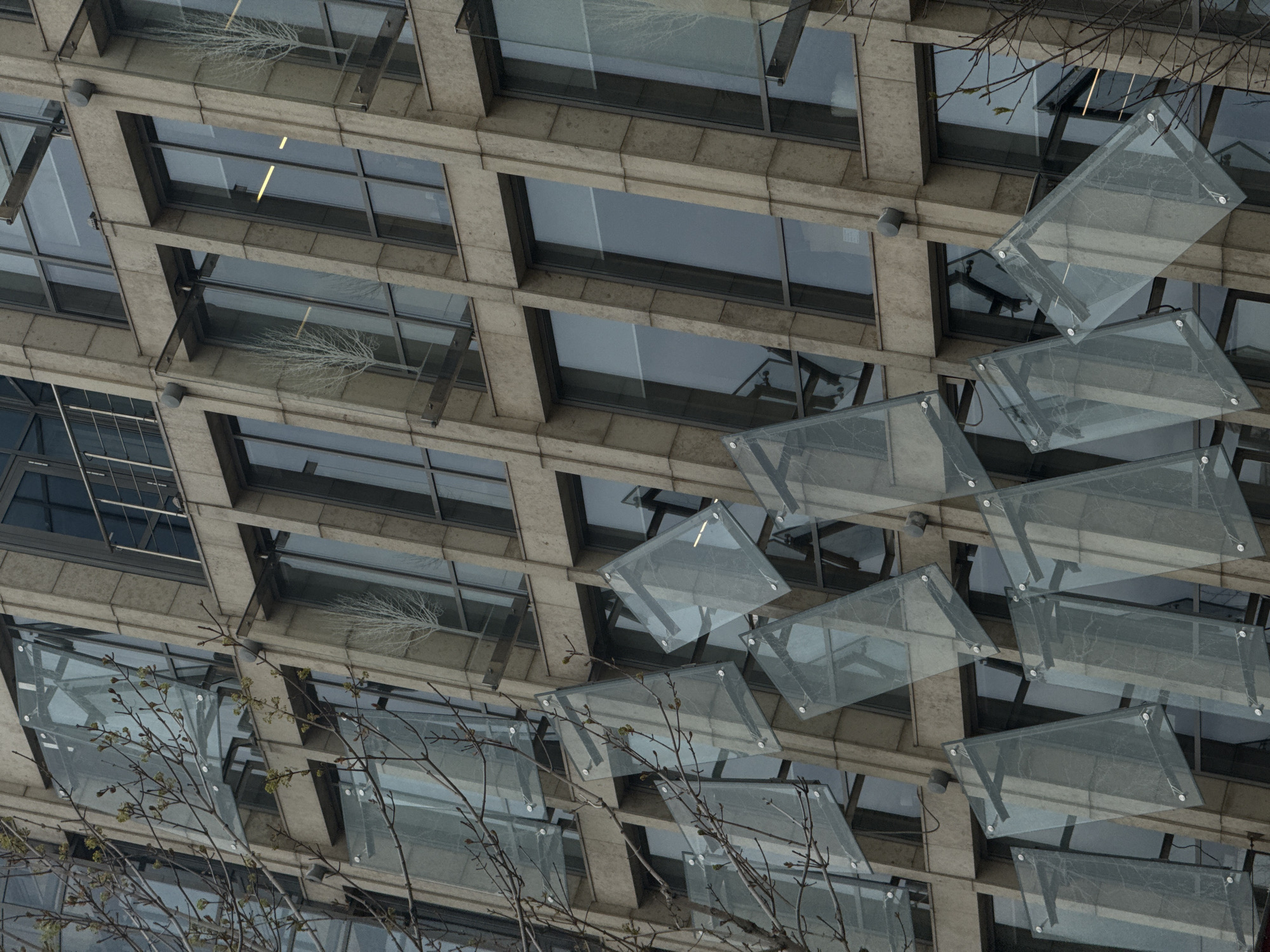 The image presents an upward-angled view of a modern building facade in Moscow, Russia, characterized by a structured grid of rectangular windows and a series of dynamic glass panels. The building's exterior appears to be constructed from light-toned concrete or stone, forming robust horizontal and vertical frames around the recessed, dark-paned windows.
A prominent feature of the facade is a repeating pattern of rectangular windows, which are predominantly dark and reflective. Several windows capture reflections of bare tree branches, integrating natural elements into the urban setting. One window clearly reflects a long, linear artificial light source from the building's interior. Small, circular, grey light fixtures are visible mounted on the building's surface beneath some window frames.
Extending diagonally across the mid-right and lower sections of the image are multiple translucent, rectangular glass panels. These panels, possibly decorative awnings or screens, are attached to the building with visible metallic fasteners and overlap, creating a layered effect and adding a sense of depth and movement to the composition.
In the foreground, particularly on the lower left and upper right, bare tree branches, some with small buds, partially obscure the building. Their stark forms create a textural contrast with the architectural lines. The overall lighting is soft and diffused, suggesting an overcast day. The color palette is muted, dominated by shades of grey, blue, and subtle greens, contributing to a somewhat cool and sophisticated aesthetic. No visible text is present in the image.
