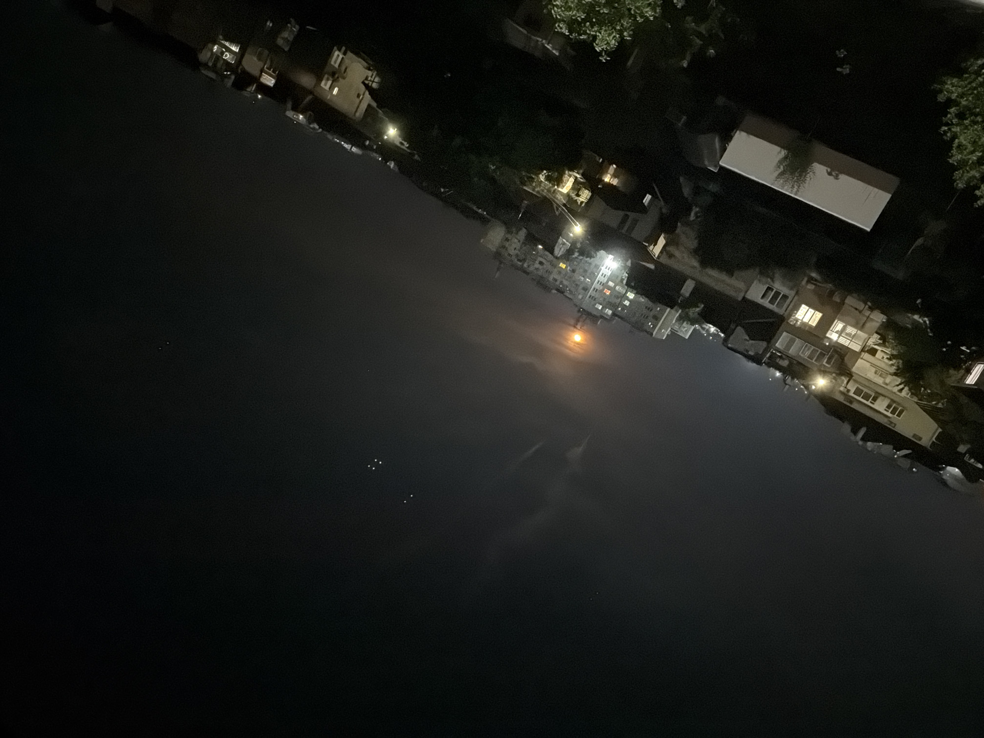 This is a nocturnal photograph showcasing an urban landscape, likely in Burgas, Bulgaria, viewed across a body of calm water. The image is significantly tilted, with the horizon line running diagonally from the lower left to the upper right.
The lower and left portion of the frame is dominated by a large expanse of dark, still water, which reflects the lights from the distant shore. A small cluster of faint white dots, possibly reflections of stars or very distant lights, is visible in the far lower left on the water's surface.
Along the upper and right side of the image, a diverse array of buildings lines the distant shore. These structures vary in size and architectural style, with many displaying illuminated windows, suggesting active occupancy or lights left on. Streetlights are scattered throughout the cityscape, casting bright points of light and illuminating sections of the buildings and surrounding environment. Dark foliage and trees are visible, interspersed among the buildings and along the waterfront.
In the mid-distance, a prominent, warm orange light source creates a hazy glow in the night sky and a corresponding reflection on the water. This could be a moonrise/moonset or a powerful artificial light. The sky is dark, indicative of nighttime, with some faint, wispy cloud formations.
The scene depicts a tranquil, illuminated urban setting at night. No specific activity or individuals are clearly visible, and there is no discernible text in the image. The overall impression is one of peaceful observation of a city after dark.