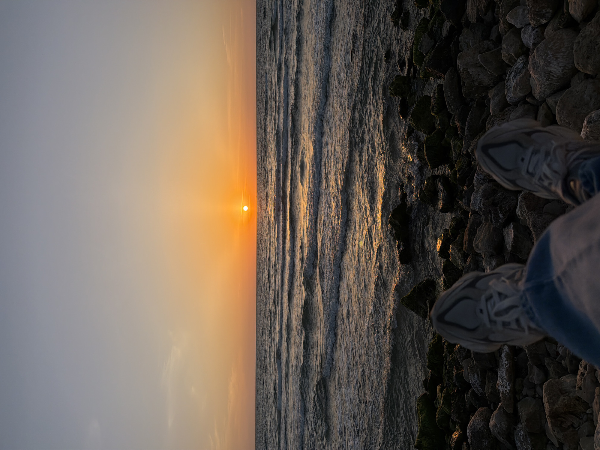 The image shows a view of the Mediterranean Sea at sunset from the shoreline in Tel-Aviv, Israel. The sky transitions from light gray to orange as the sun sets over the horizon. The sea is rough with waves reflecting the orange glow of the sun. On the right side of the image, there is a rocky shoreline. A pair of sneakers is visible, belonging to the person taking the picture. The lighting is dim, casting shadows on the rocks.