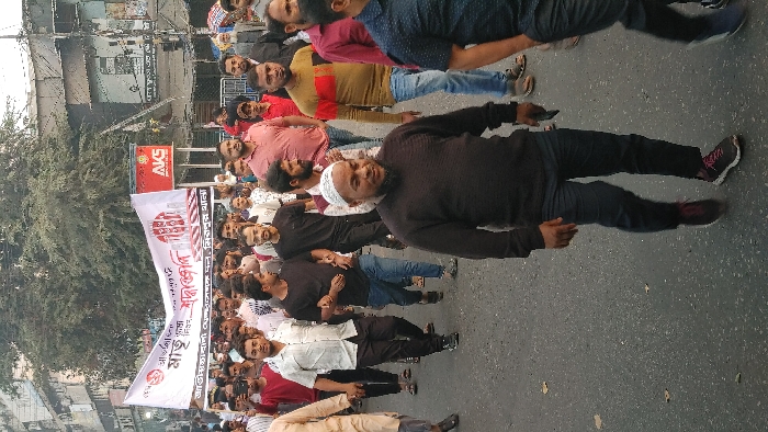 A street scene with a large group of people lying down in the road. They appear to be protesting, as they are holding a large banner with writing on it. Most are dressed casually in dark clothing. The man in the front is wearing a black shirt, black pants and a white kufi. In the background, buildings and trees line the street. The road is grey and dusty, indicating a dry climate.