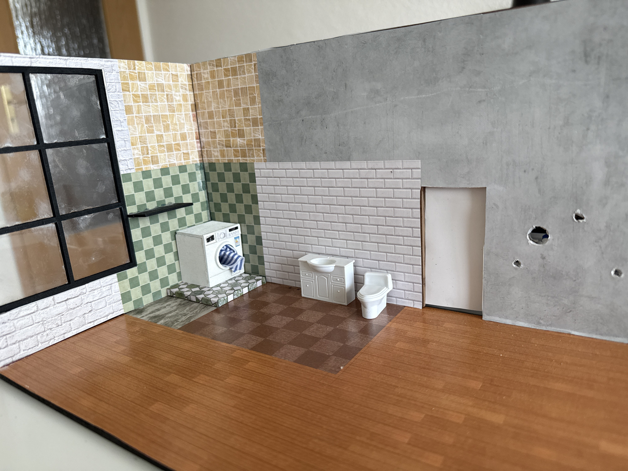 This is a close-up, high-angle, eye-level shot of a miniature diorama depicting a bathroom scene. The diorama has a wooden floor with a warm, orange-brown tone. In the background, walls are decorated with various tile patterns: a white brick pattern on the left, a green and white checkered pattern above that, and a yellow tiled wall to the right of the green tiles. A large window with black mullions and clear panes is positioned on the far left, looking into a space with a reflective surface.
In the center of the diorama, a white washing machine with a blue and white striped towel inside is placed on a tiled platform. Adjacent to the washing machine is a white vanity with a sink and a white toilet. These fixtures are against a wall covered in white subway tiles. To the right of the subway tiles is a larger wall section that appears to be a light gray concrete texture, with a doorway cut into it. The concrete wall has two visible circular holes and one smaller hole.
The floor in the bathroom area has two distinct patterns: a section with brown and lighter brown checkered tiles, and a section with a marbled gray and green pattern beneath the washing machine. The diorama appears to be an indoor scene and does not provide any context or details related to Lutherstadt Eisleben, Germany.