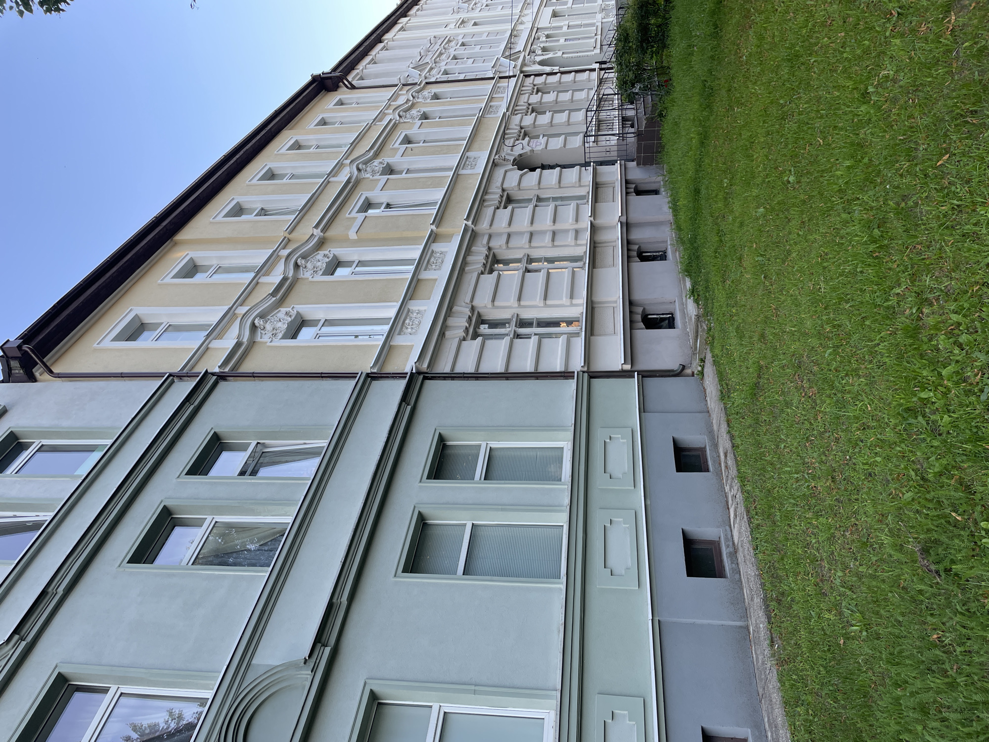 The image shows a side view of a row of connected apartment buildings in Sovetsk, Russia, under a clear blue sky. The buildings are constructed with distinct architectural styles and colors. The closest building on the left has a pale green facade with grey detailing near the bottom and around the windows. It features multiple windows, some with blinds, and decorative architectural elements. Adjacent to it, and extending into the distance, are two buildings with creamy yellow facades and white decorative trim around the windows and along the levels. These buildings exhibit more ornate detailing, including sculpted elements above some of the windows. The ground in front of the buildings is covered with a lush green lawn. The scene appears to be a residential area, captured in what appears to be late afternoon or early evening light.