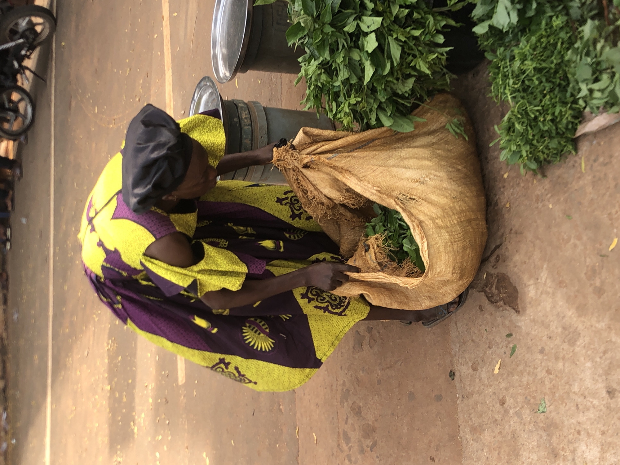 This image captures a woman seated on the ground outdoors, likely within a market or roadside setting in Nsugbe, Nigeria, under bright daylight.
The main subject is a woman, seen from an elevated perspective, wearing a vibrant traditional wrapper dress with prominent yellow and purple patterns, and a black head covering. She is leaning over a large, light brown woven bag filled with fresh green leafy vegetables, with her hands inside the bag, suggesting she is sorting, arranging, or preparing the produce.
Surrounding her are several large piles of the same type of green leafy vegetables, indicating a considerable quantity of produce, possibly for sale. Behind the woman, two large, dark-colored metal drums or containers, each topped with a metal lid, are visible. To the far left of the frame, the front wheel and part of the engine of a motorcycle are partially visible, suggesting a busy environment or a means of transport. The ground beneath her is a mix of compacted dirt and concrete.
The scene conveys a sense of daily life and commerce in an outdoor market or roadside stall. There is no visible text in the image.
