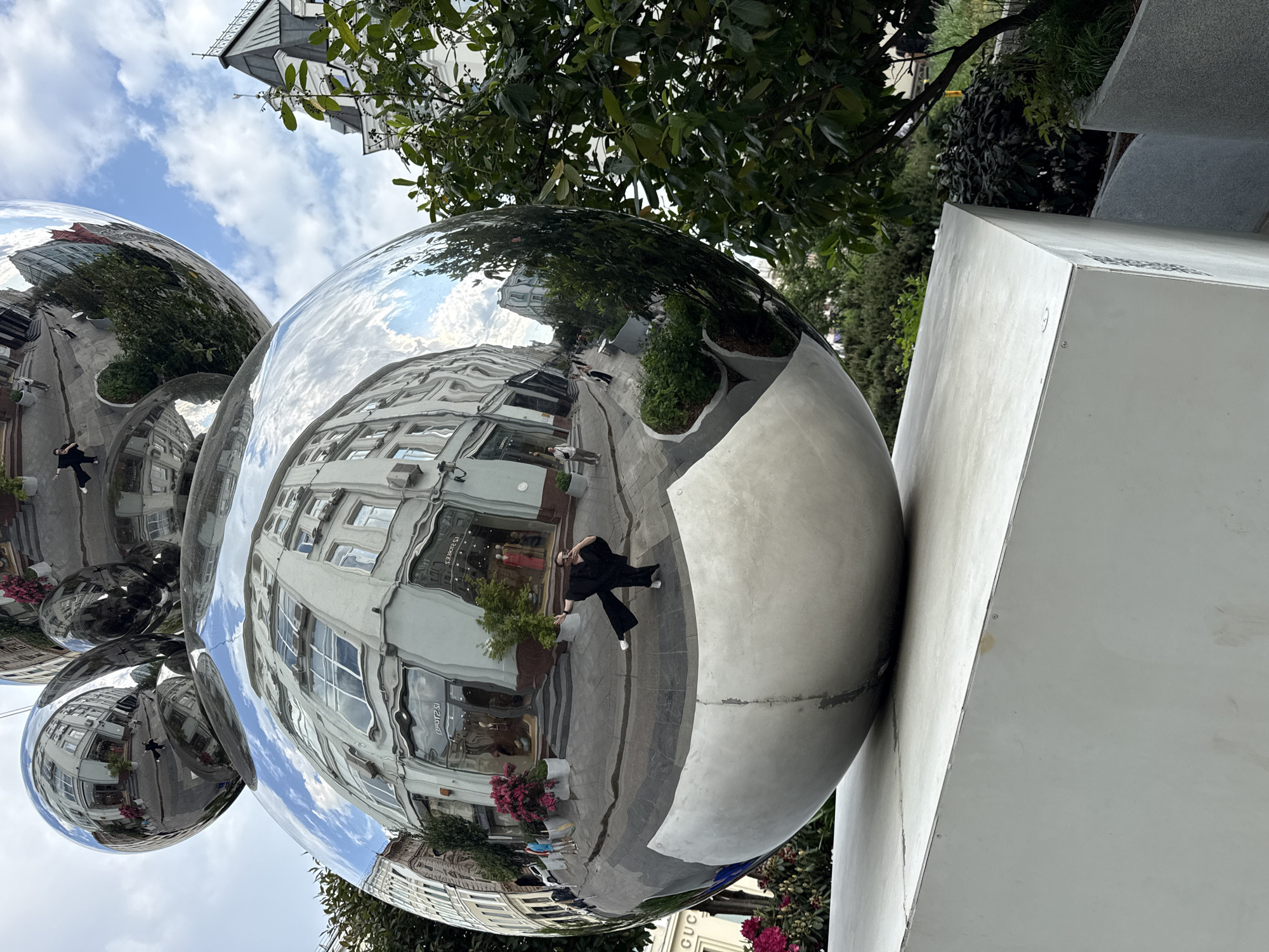 A distorted and inverted image of a city street scene reflected in large, polished, spherical metallic sculptures. The largest sphere is the most prominent, reflecting buildings, sidewalk, and foliage. In the reflection of the sphere, a person dressed in black stands on the sidewalk. The spherical structure appears to be a cluster of interconnected orbs. These sculptures are situated adjacent to a large, light-grey rectangular structure. The foliage is both above and around the sculpture. The image includes the location as Moscow, Russia, based on the storefront lettering that is reflected in the sphere. The sky is visible above the sculptures, and a building is also visible behind the leaves.