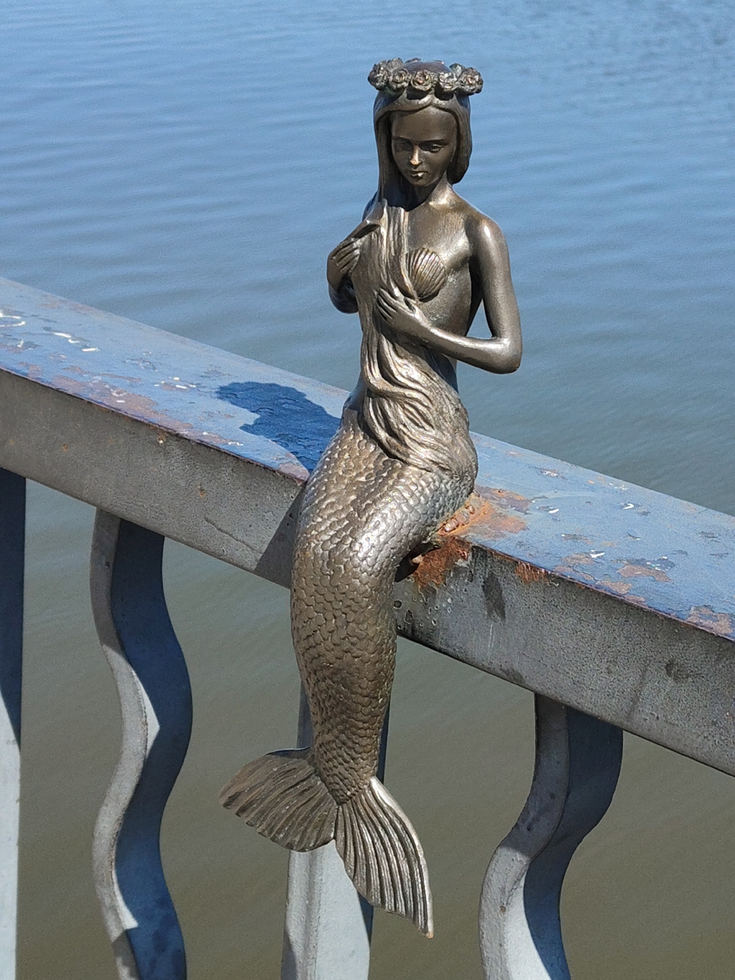 A bronze statue of a mermaid is perched on the railing of a bridge in an unknown city. The mermaid has long hair, a flower crown, and a scaly tail. She is holding her hair with both hands. The railing is blue and rusty. The background shows a body of water.