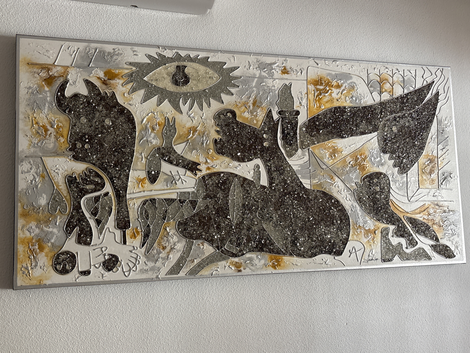 The image shows a textured, rectangular artwork with abstract figures rendered in dark, shimmering material and metallic silver and gold tones. The artwork is mounted on a white wall, which appears to be a textured plaster or stucco finish. The lighting suggests an indoor setting, likely a home or gallery, with natural light coming from an unseen source casting subtle shadows on the wall and artwork. There are no people visible in the image. The artwork itself features an eye-like symbol at the top, surrounded by a jagged edge. Below this, several stylized figures are depicted, some appearing humanoid, others more animalistic or monstrous, with sharp angles and flowing lines. The materials used create a sense of depth and shimmer, with the dark material resembling crushed glass or a similar aggregate. The overall aesthetic is modern and possibly surrealist. There is no visual information in the image to indicate its location in the city of Orihuela, Spain.