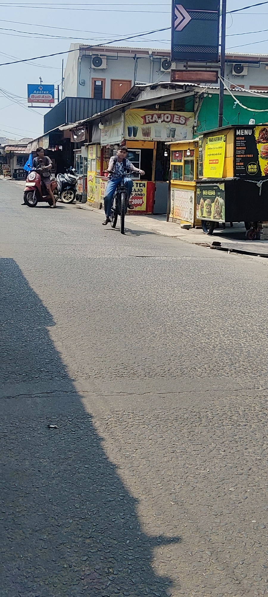 In the city of Mekarmukti, Indonesia, on a sunlit day, a scene unfolds along a paved road lined with small businesses. The asphalt surface is textured and marked with faint cracks. To the left of the frame, a shadow of an unknown object stretches across the road.
In the middle ground, a young person is riding a bicycle, wearing a patterned short-sleeved shirt, blue jeans, and a backpack. They are positioned on the right side of the road, moving away from the viewer. Further to the left, another individual is seated on a red scooter, also moving away from the viewer, wearing a dark t-shirt and shorts.
The businesses lining the road feature colorful stalls and signage. A prominent yellow stall displays the text "RAJO ES" and images of drinks. Another stall, also yellow, shows an advertisement for "BKB KEBAB" with pictures of kebabs and burgers. A menu board is visible on this stall. Adjacent to these is a stall with red and white signage, and another stall with blue and white signage. Above some of these stalls, a blue sign for "Apotek Budi Asih" is mounted. The buildings behind the stalls appear to be multi-story structures with visible air conditioning units. Overhead, electrical wires are strung across the sky, and a directional sign with arrows is positioned on a pole.