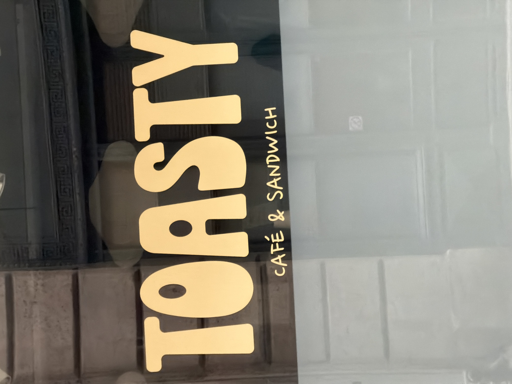 The image depicts a window display of "TOASTY CAFE & SANDWICH", a cafe located in Paris, France. The letters are in gold and vertically oriented. The background is a blurred reflection of the street and a building with an ornamental facade. The lighting is soft and diffused, with a noticeable shift in light across the window's surface. The focus of the image is on the shop's name and its offering.