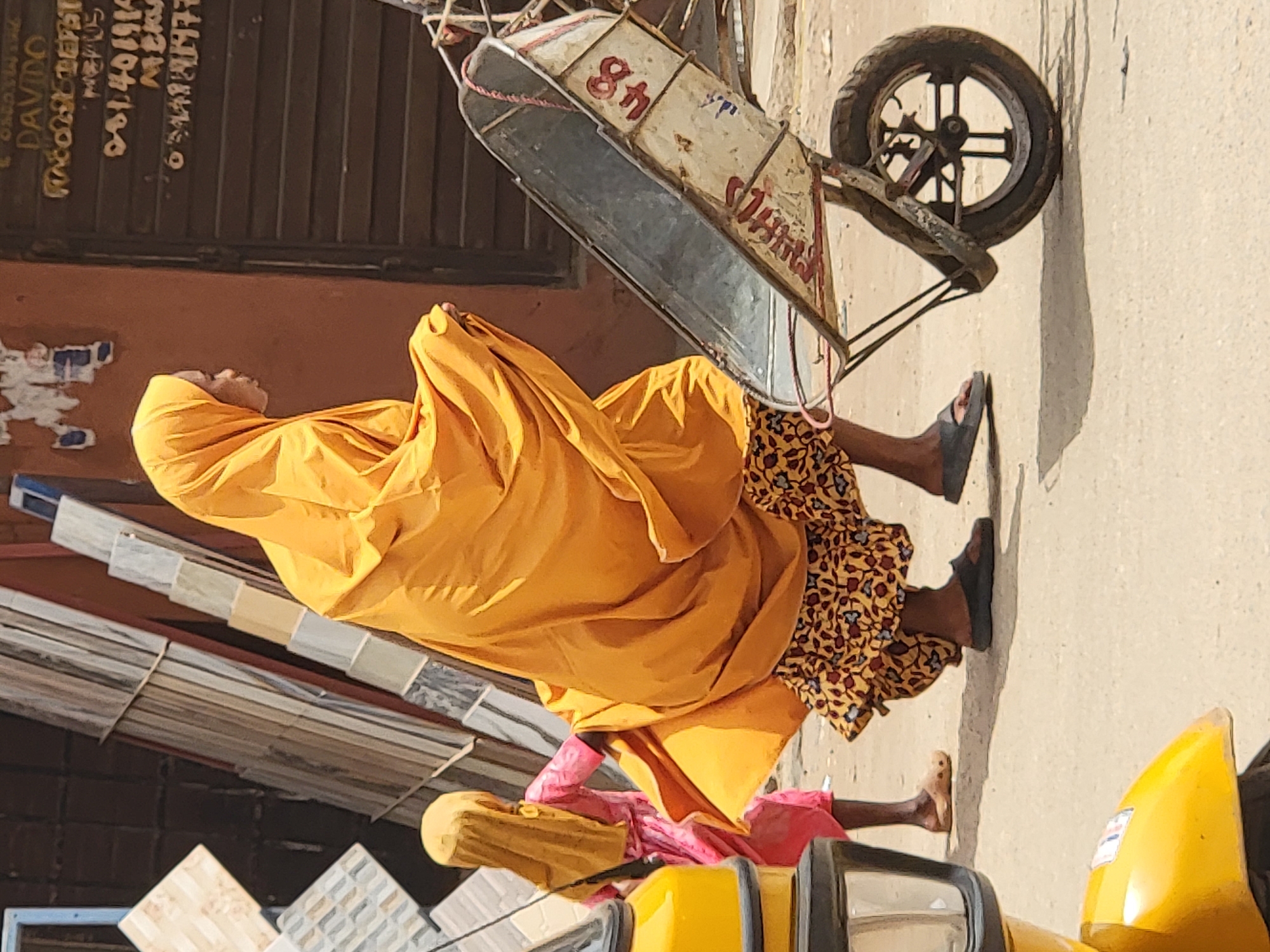 A bright, sunlit outdoor scene in Maiburiji, Nigeria, captures a person and a child walking or standing near a street. The image is captured in daylight, casting distinct shadows, such as the one beneath a wheelbarrow.
The main subject is an adult, seen from behind and slightly to the side, wearing a flowing, vivid yellow-orange garment that covers their head and most of their body. A patterned skirt, featuring a yellow, brown, and black floral or geometric design, is visible beneath the main garment. The person wears dark-colored sandals. Partially visible behind the adult is a child, wearing a pink garment and possibly a yellow head covering, with only their legs and lower torso clearly seen.
To the right, an old, metal wheelbarrow with a single spoked wheel is present. Its basin is a mix of grey metal and white patches, on which the red numbers "84" are clearly visible, along with other indistinct red markings that might spell "WINA". A red rope or cord is tied around the wheelbarrow frame.
In the background, on the left, is a building or wall. The upper section appears to be made of dark brown, possibly corrugated metal or wooden panels. Below this, there's a lighter brown/orange wall with some white, peeling paint or residue. White, rotated text is visible on the dark brown section, including "DAVDO", "08140191", and fragments like "ME". Stacked tiles of various light colors (white, grey, beige, marbled) are arranged on the lower left, suggesting a tile display or shop. A portion of a yellow vehicle, possibly a tricycle or motorcycle taxi, is visible in the bottom right corner of the frame.