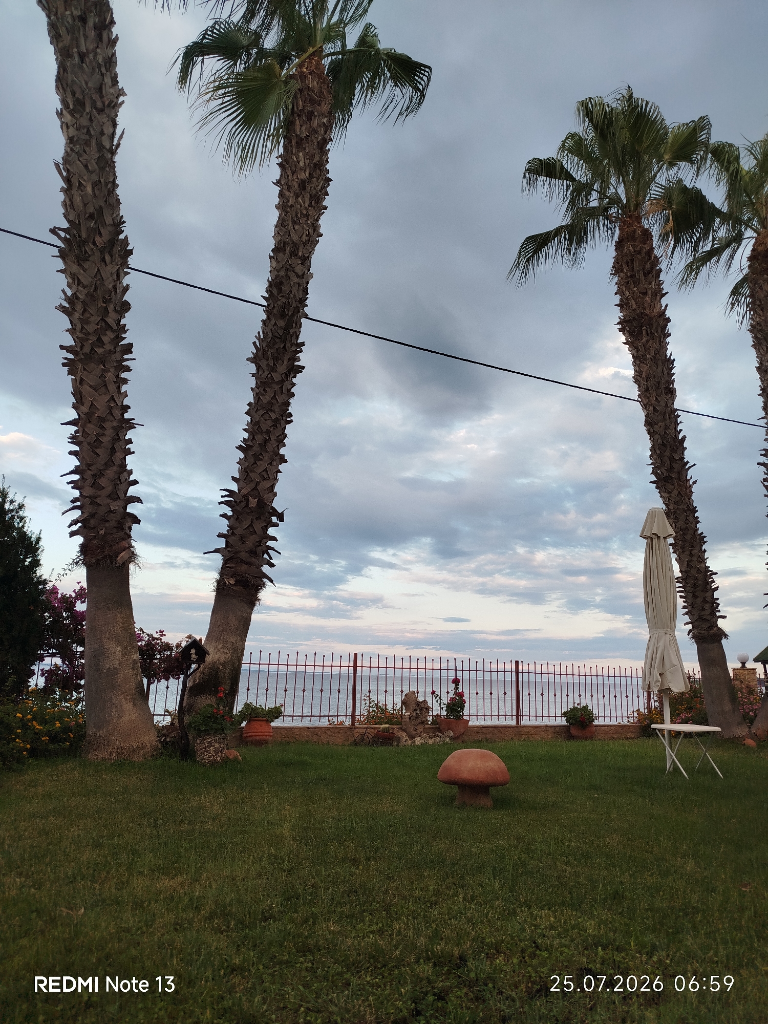This image depicts an outdoor scene with several palm trees in the foreground, silhouetted against a cloudy sky. A body of water, likely the sea, is visible in the background, separated from the foreground by a decorative metal fence. The lawn in the foreground is green and well-maintained, with a small mushroom-shaped ornament in the center. A folded white umbrella and a small white table are also present on the lawn. The image is dated 25.07.2026 06:59, and the text "REDMI Note 13" is in the bottom left corner. The location is identified as Sithonia Municipal Unit, Greece. There are no people or animals visible in the image. The overall atmosphere is peaceful and serene, suggesting a seaside resort or a private garden.