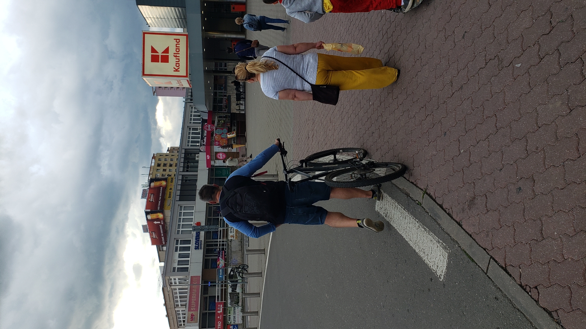 The image depicts a paved outdoor area in front of commercial buildings. A man in a blue long-sleeved shirt and denim shorts is walking with a bicycle, pushing it alongside him. He is wearing a backpack. Behind him, a woman in a striped t-shirt and yellow pants walks away from the camera, carrying a plastic bag. Further in the background, other pedestrians are visible. The buildings have various signs, including "COOP SUPERMARKET," "SLOVENS," and a prominent "Kaufland" logo. The environment suggests a pedestrian walkway adjacent to a road, with overcast skies overhead. This scene is located in Prešov, Slovakia, specifically in front of the Kaufland supermarket on Armádneho generála Svobodu street, where the Coop supermarket is also located.
