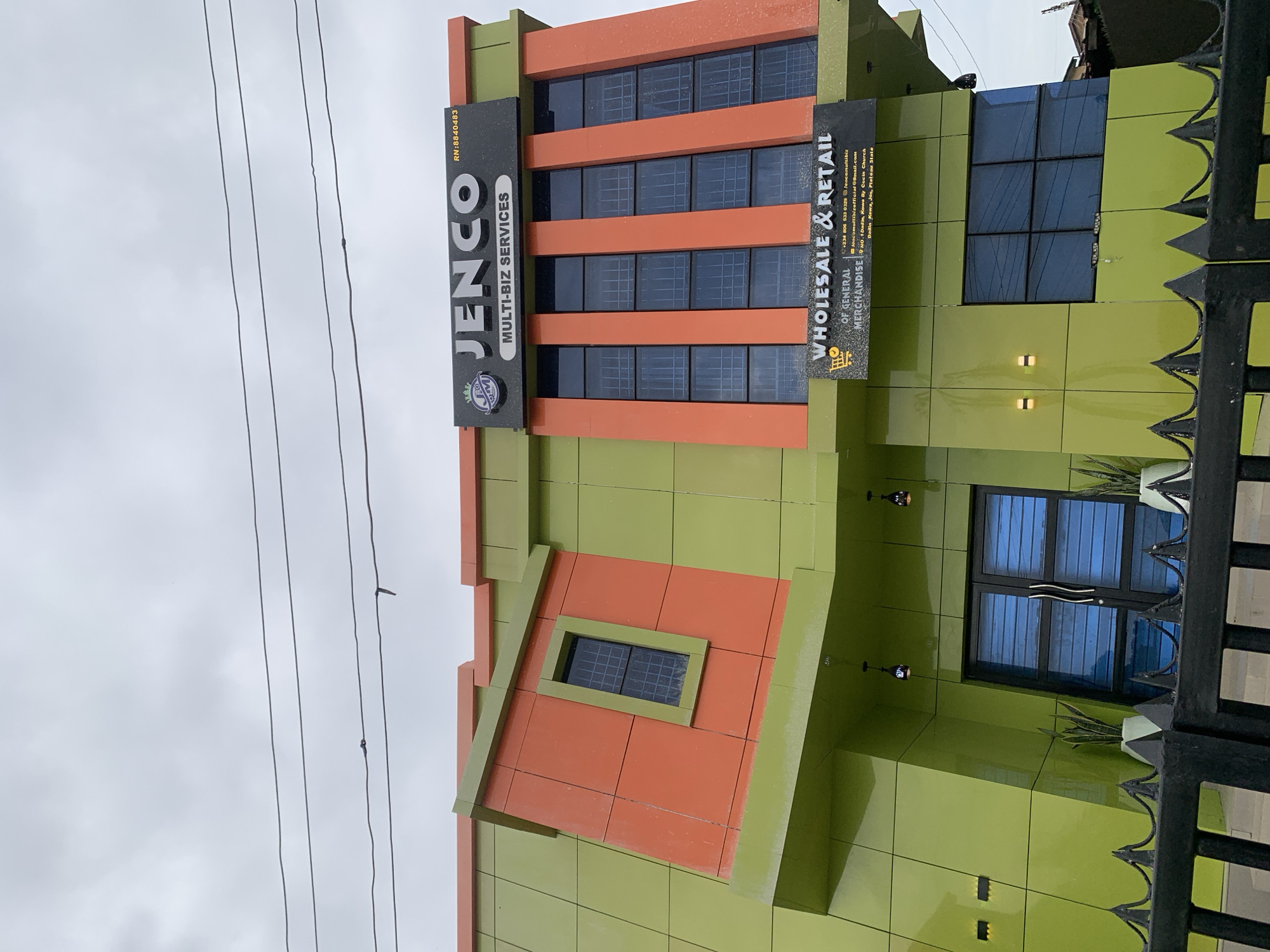 An exterior view captures a two-story commercial building, identified as "JENCO MULTI-BIZ SERVICES," under an overcast sky. The building features a striking lime green and orange color scheme. The upper left section presents an orange panel set at an angle, containing a single rectangular window. The upper right facade is orange with five vertical rectangular windows. A black sign atop the building displays "JENCO MULTI-BIZ SERVICES" with a crown logo and "RN: 8840483." A second black sign below the upper windows announces "WHOLESALE & RETAIL OF GENERAL MERCHANDISE" alongside a shopping cart icon. This sign provides contact details: phone number "+234 806 532 0329," Instagram handle "@jencomultibiz," email "jencomultibizofficial@gmail.com," and the physical address "NO. 1 Dadin Kowa, By Cochin Church Dadin Kowa, Jos, Plateau State."
The ground floor, primarily lime green, includes a double-door entrance with blue-tinted panels, flanked by two potted snake plants. Two black pendant lights hang above the entrance, and two horizontal wall-mounted lights are visible to the left. A large rectangular window is situated to the right of the entrance. Multiple black power lines traverse the upper portion of the image against the gray sky. A black metal fence with pointed finials partially obstructs the lower foreground of the building. No individuals or specific actions are observed within the frame.