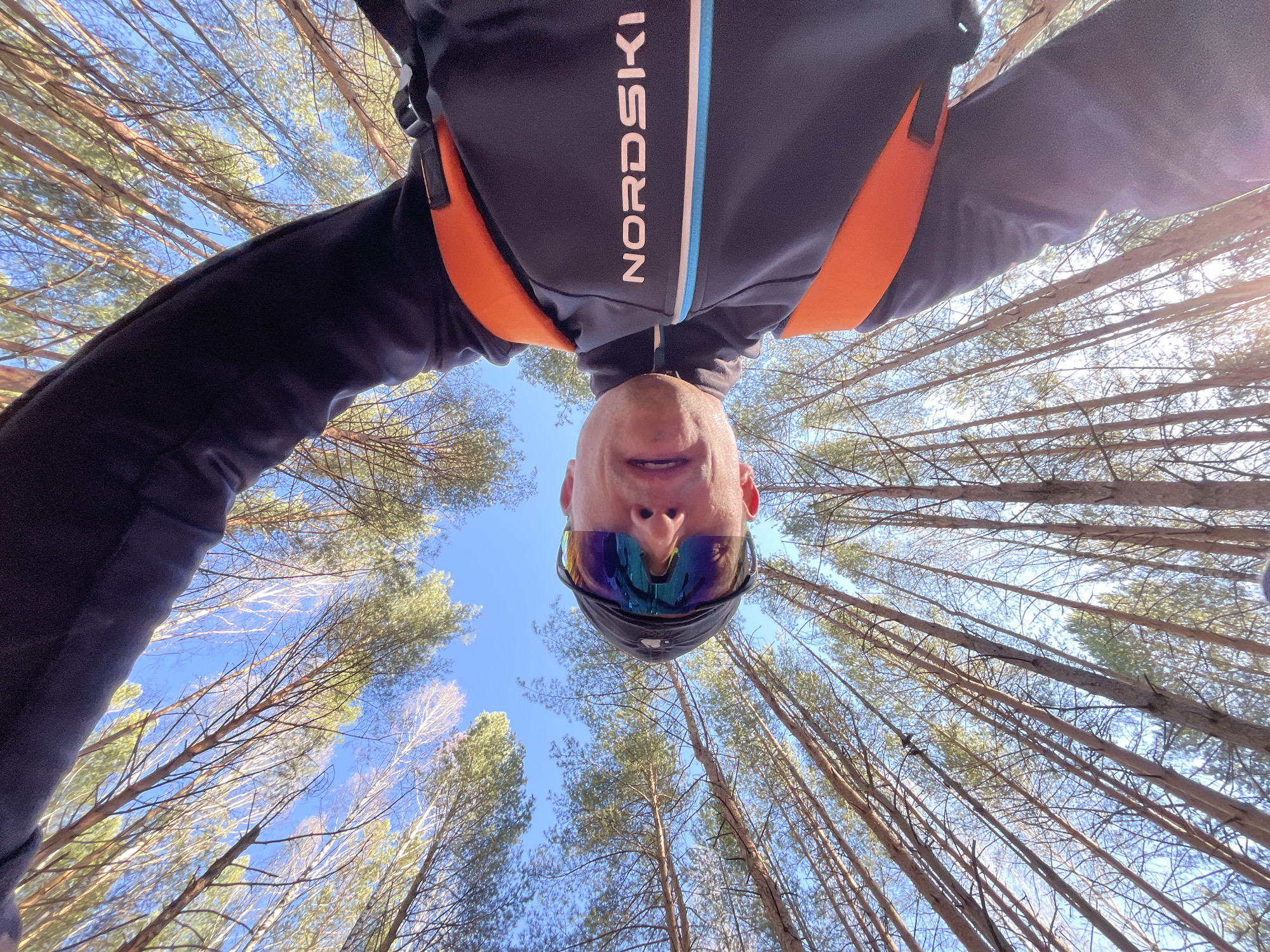 A man wearing a dark "NORDSKI" branded jacket, cap, and mirrored sunglasses, is pictured from a low angle, looking directly down at the camera. The background is a forest, with tall, slender trees stretching towards the blue sky. The man's face is centered in the frame, and the trees form a circular pattern around him, creating a sense of depth and perspective. The location is Chaikovsky, Russia.