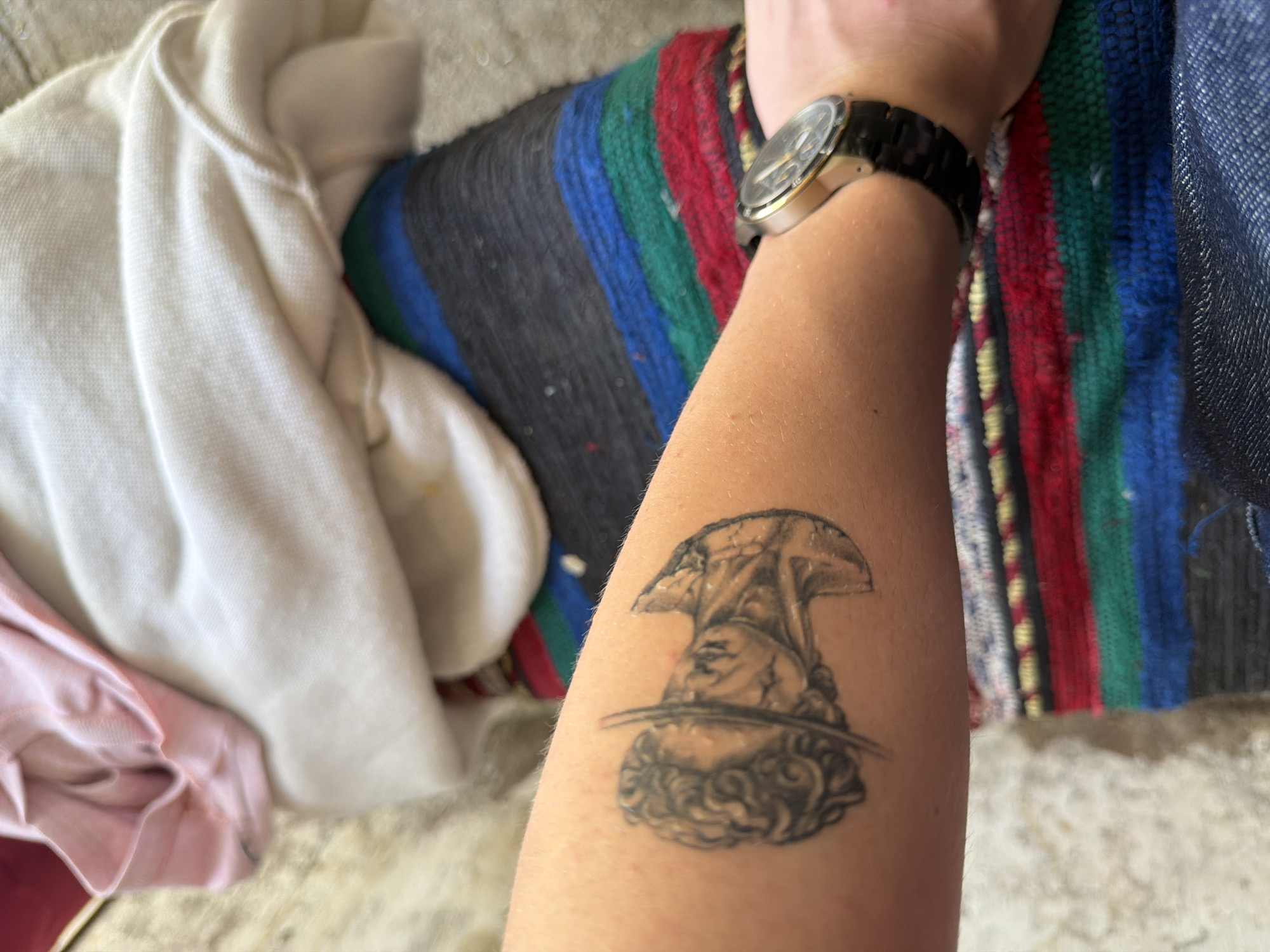 A close-up shot shows a forearm with a tattoo of an upside-down bust of a man, possibly a classical sculpture, positioned on the lower part of the arm. A gold-toned wristwatch with a black band encircles the wrist. Adjacent to the arm and tattoo are various items, including a light-colored, possibly white or cream-colored garment or fabric and a colorful striped blanket. The environment seems to be indoors. The image does not show the city of Kanchevo, Bulgaria, or any specific location. There are no people or actions depicted besides the presence of an arm with a tattoo.