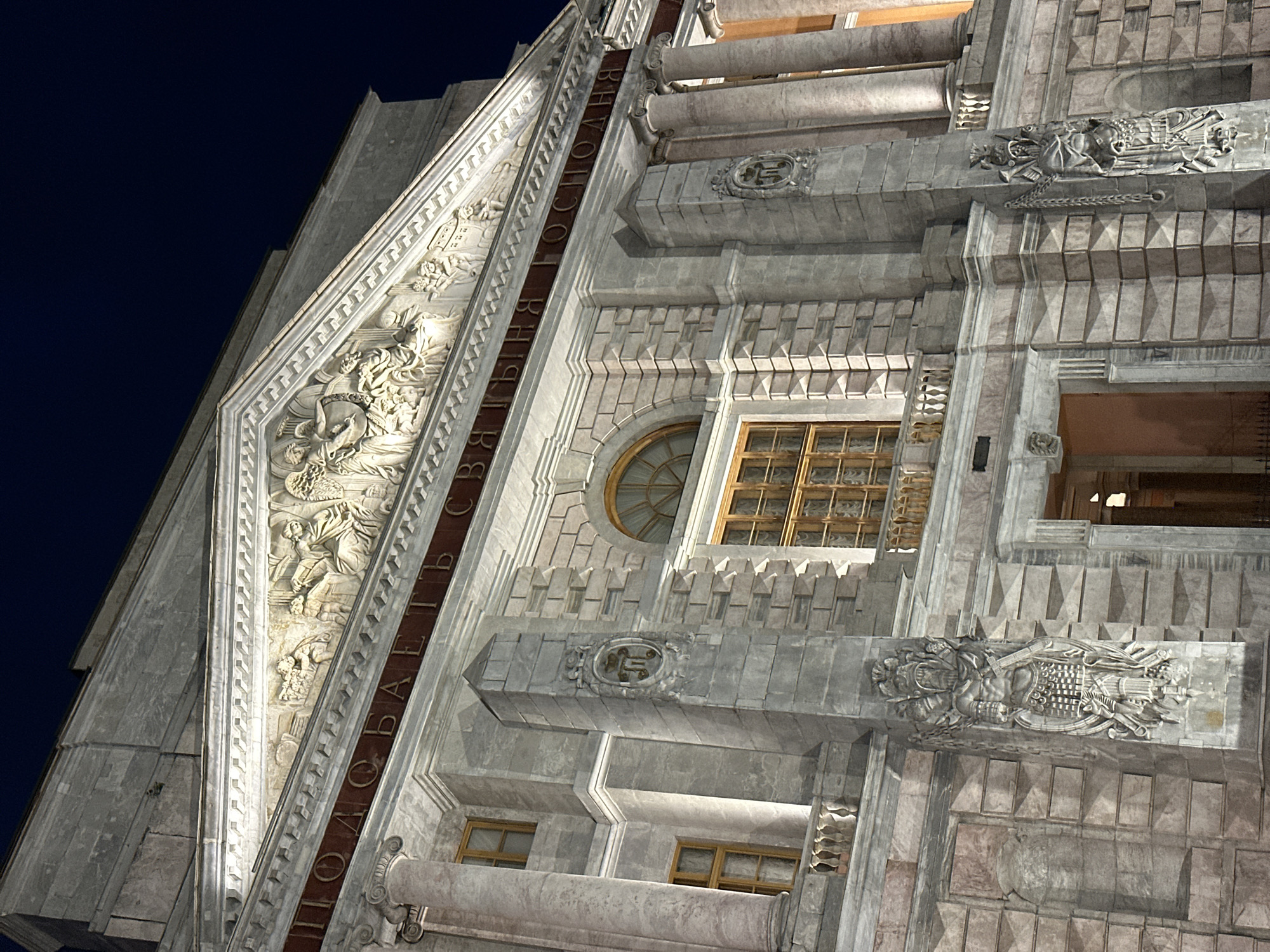 The image shows a close-up view of the facade of the Winter Palace in St. Petersburg, Russia, at night. The palace, illuminated by artificial lights, displays its grand architectural features. The focus is on the upper section of the building, showcasing intricate bas-relief sculptures adorning the pediment. These sculptures depict figures engaged in various activities, possibly representing historical or allegorical scenes. Below the pediment, a band of dark red with gilded Cyrillic lettering runs across the facade. The stonework of the building is detailed, with classical columns and arched windows visible. Two monumental sculptures, resembling warriors or mythical figures in armor, flank the central entrance area. The sky above is a deep, dark blue, indicating nighttime.