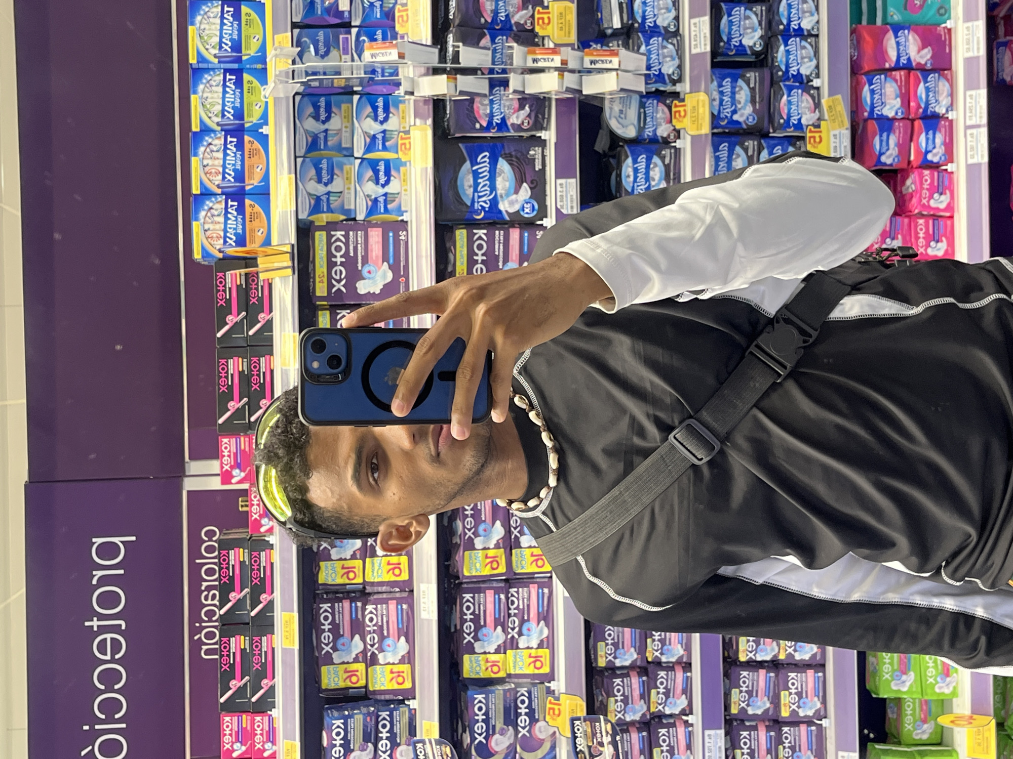 A man is taking a selfie in an aisle of a store. The aisle is filled with feminine hygiene products, mainly pads, from brands like Kotex and Always. The man is holding a blue iPhone with a black case. He is wearing a black shirt with white sleeves and a white trim, along with a shell necklace. A black bag strap is across his chest. The location is not clear from the provided image and text.