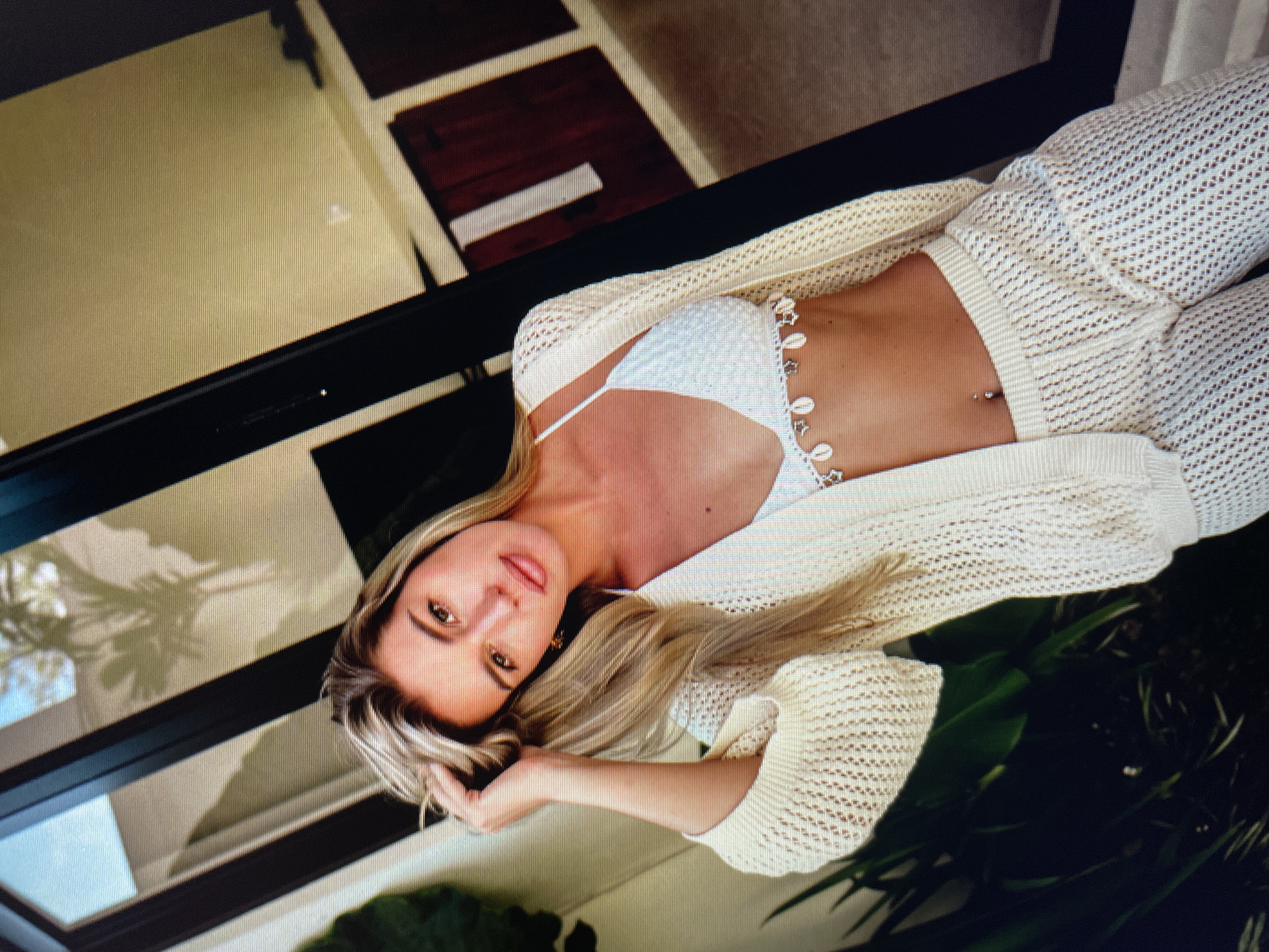 A woman with blonde hair and tanned skin is posing for a photo. She is wearing a white knitted bikini top with seashells and a white knitted cardigan and pants. Her left hand is in her hair, and she is looking at the camera. The background appears to be an outdoor setting, possibly a patio or balcony, with plants and glass doors. The lighting suggests it is daytime. The setting is consistent with a location in North Kuta, Indonesia.