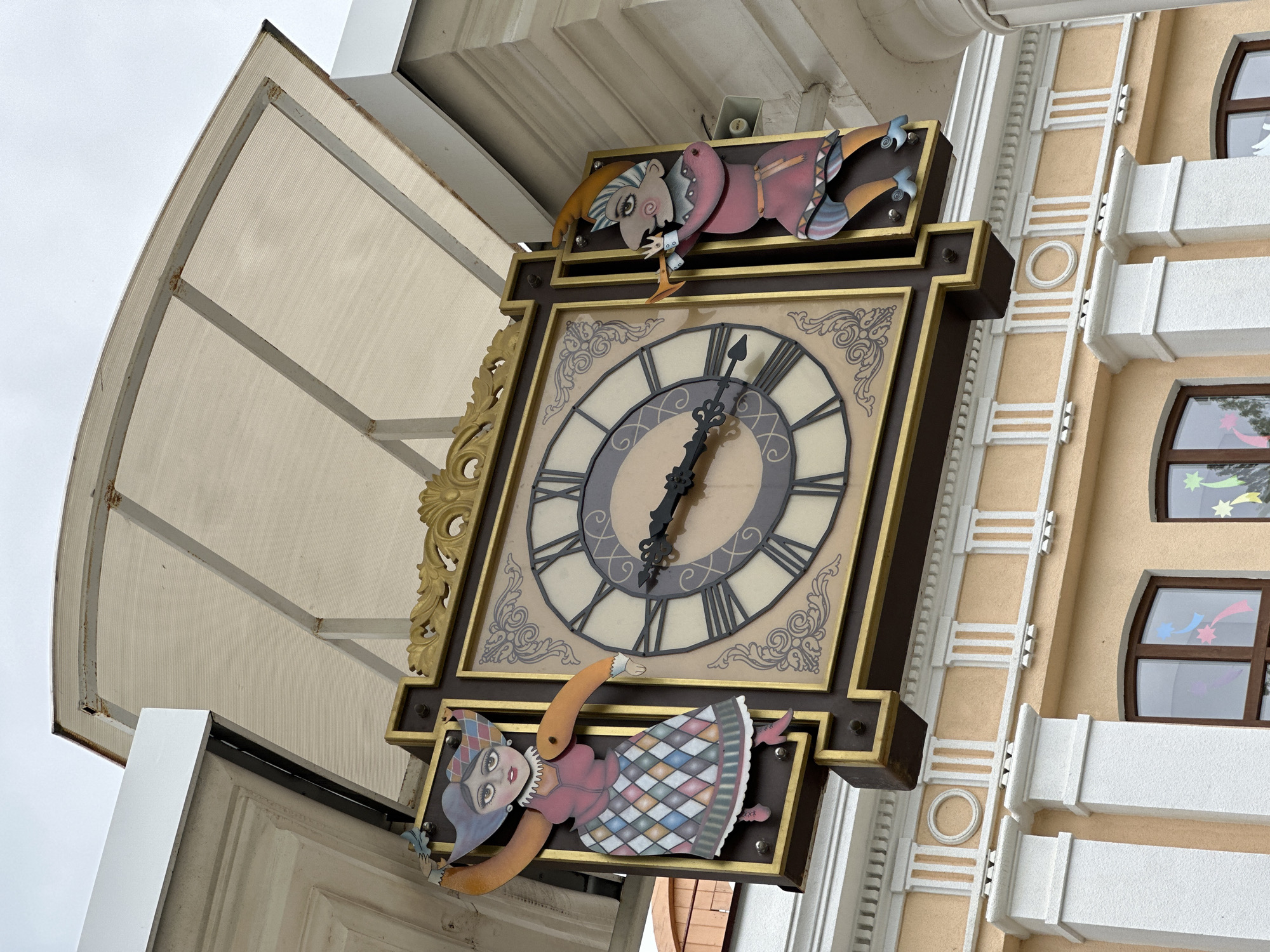 The image shows a large, ornate clock mounted on a building in Stavropol, Russia. The clock has a brown frame with gold accents and Roman numerals. Two whimsical figures, one resembling a jester and the other a gnome-like character, flank the top and bottom of the clock. The building itself is peach-colored with white trim and windows that appear to have decorative star decals. The architectural style suggests a historical or themed district. Above the clock, a white canopy structure provides a partial covering. The time displayed on the clock is approximately 6:02.