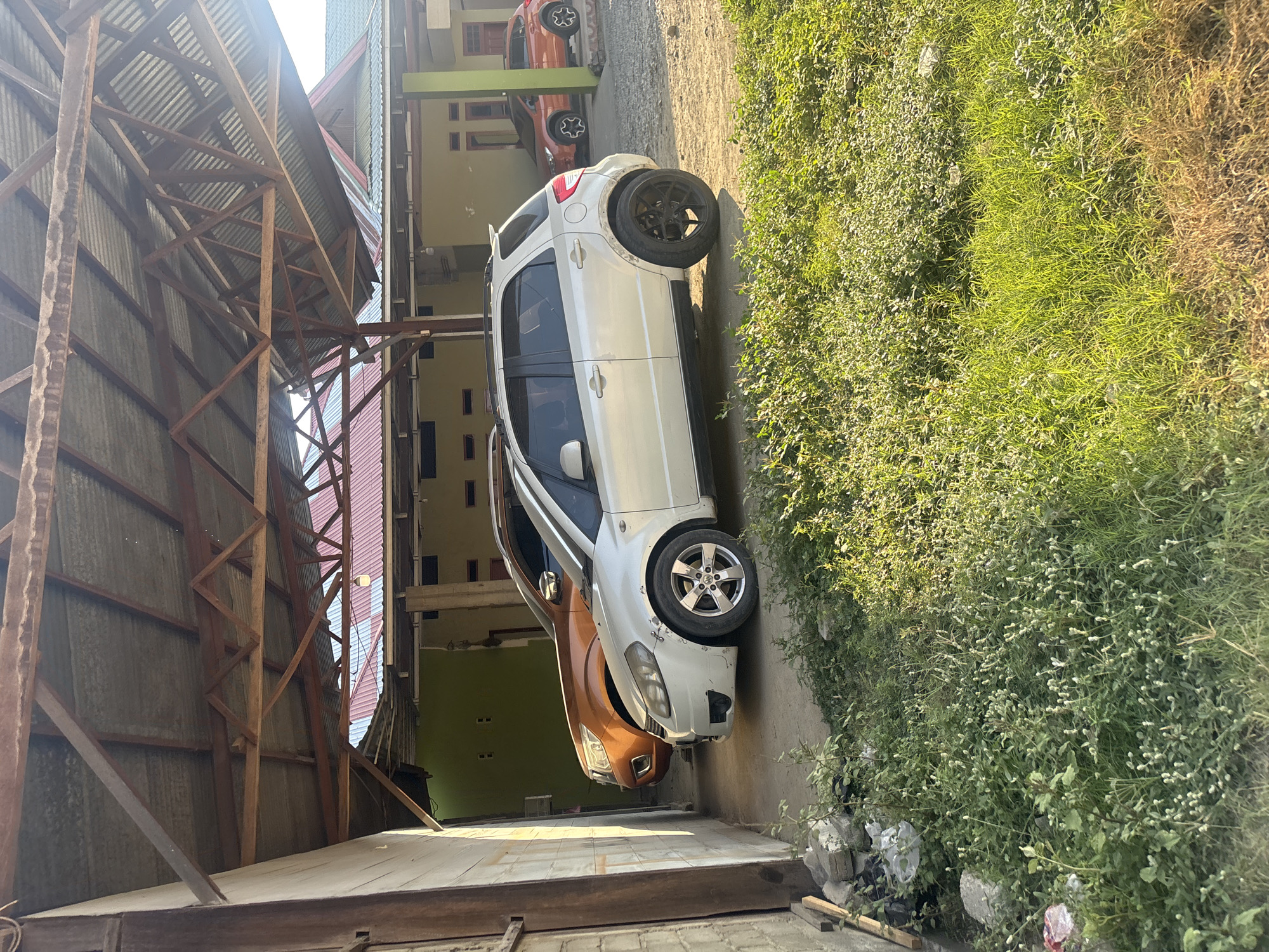 The image shows two cars parked under a covered structure. The structure has a corrugated metal roof supported by wooden beams. The car in the foreground is a white hatchback with black rims on the rear wheels and silver rims on the front wheels. It appears to have some body damage on the rear fender. The car behind it is an orange SUV. In the background, a yellow building with a red-roofed addition is visible. The area is overgrown with grass and weeds. The scene appears to be located in a residential area in Bunto, Indonesia, likely a driveway or carport. The lighting suggests it is late afternoon.