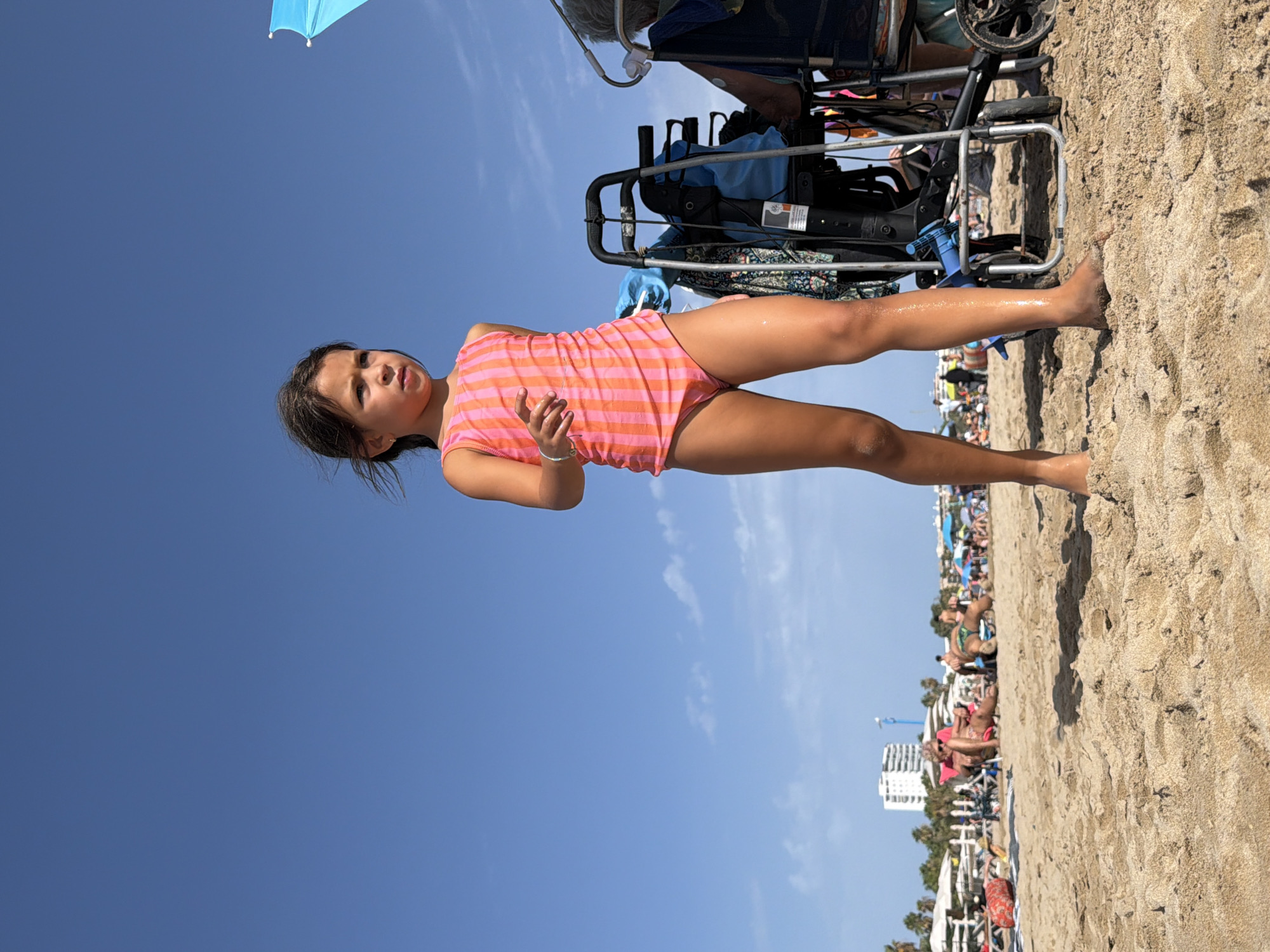 A young girl with dark hair, wearing a pink and orange striped swimsuit, stands on a sandy beach in Salou, Spain. She is looking to her left. The sky above is clear and blue, with a few wispy clouds. In the background, other beachgoers are visible, some lounging on chairs and others walking along the shore. Beach umbrellas and tents dot the landscape. Behind the girl, a blue umbrella is partially visible, and to her right, a wheeled cart and a person in a wheelchair are present. Buildings and palm trees can be seen in the distance. The sand is light brown and textured.