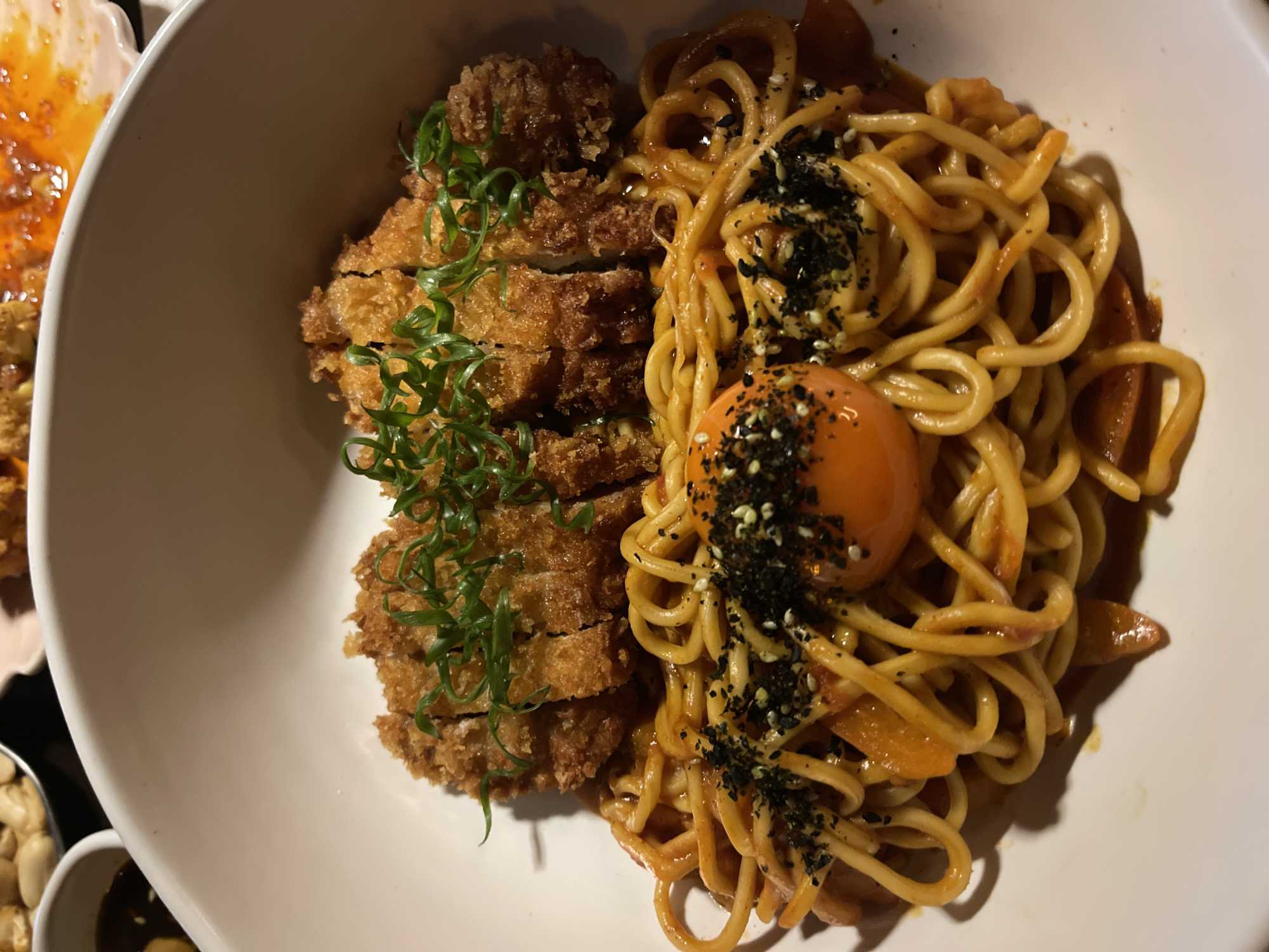 A close-up, overhead shot captures a white bowl filled with a Japanese-style dish. The bowl is divided into two distinct sections. On the left, sliced, golden-brown breaded cutlets are arranged vertically, garnished with vibrant green scallions. To the right, a generous portion of thick, yellow noodles is bathed in a reddish-orange sauce, sprinkled with black and white sesame seeds. A bright orange egg yolk sits atop the noodles, adding a rich focal point.
The lighting suggests an indoor setting, possibly a restaurant, and the deep shadows indicate the time of day is likely evening or night. The food appears to be freshly prepared and ready to be eaten, creating a sense of immediate enjoyment. The presentation is neat and appetizing, with the textures of the crispy cutlets and smooth noodles contrasting visually. No text is visible in the image. The context provided is Mengwi, Indonesia, hinting at the culinary origins or location of this meal.