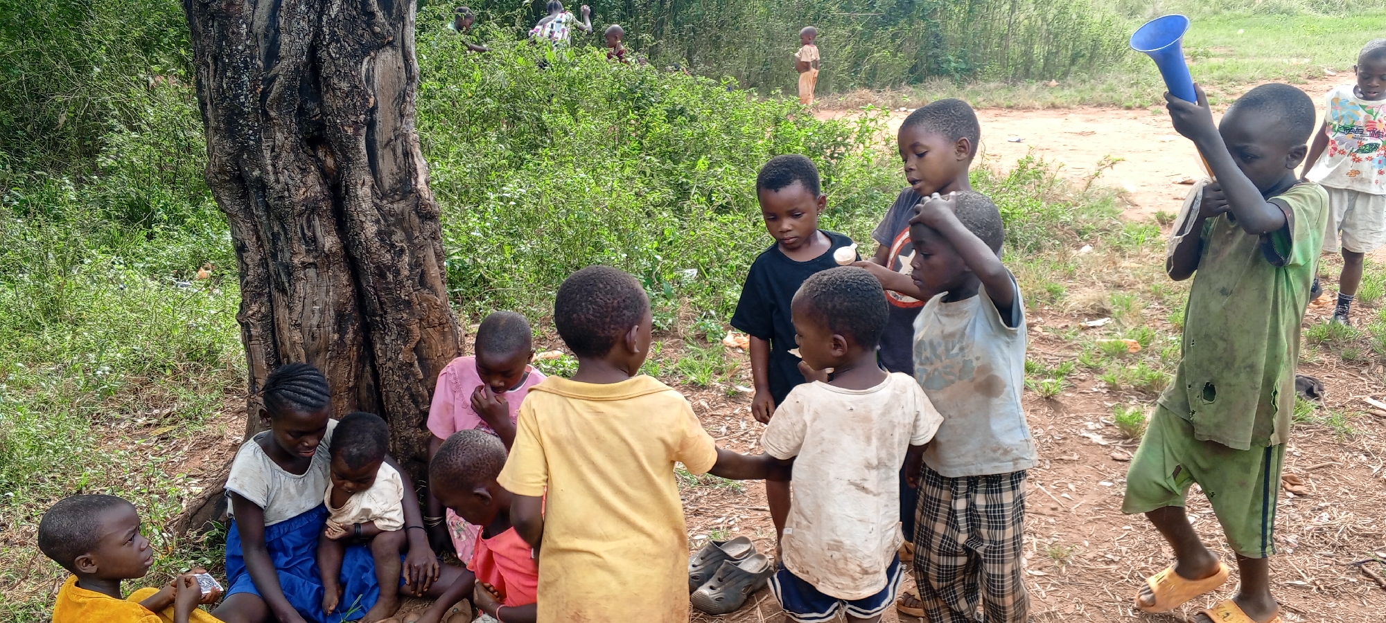 This outdoor image captures a diverse group of approximately fifteen children, ranging from toddlers to pre-teens, in Ngalula, Democratic Republic of the Congo. The scene unfolds on a dirt ground, framed by a large tree trunk on the left and dense green foliage in the background, suggesting a rural or semi-rural setting.
Several children are gathered near the base of the tree. An older girl in a blue dress and white top is seated, holding a baby or toddler. Other children in various simple shirts and dresses, including one in pink and another in yellow, are seated or standing nearby. Towards the center and right, a cluster of boys is visible. One boy wears a black t-shirt featuring a red, white, and blue shield logo, likely Captain America's, with faint, illegible text beneath it. Another boy in a light-colored, possibly plaid, t-shirt holds a small white spoon. To the far right, a boy in a worn, ripped green t-shirt and shorts holds a bright blue funnel-shaped object aloft, possibly playing with it as a toy or megaphone. Next to him, another boy wears a white t-shirt with stylized text, where "TIME" is clearly visible. A pair of black sandals rests on the ground near the central group, and the boy with the blue funnel wears light brown sandals.
The children appear to be engaged in informal play or simply socializing. Their expressions are calm and focused, with no overt display of strong emotions. The lighting suggests it is daytime, possibly late morning or afternoon, under clear or partly cloudy skies. The overall atmosphere depicts a slice of daily life in the region.