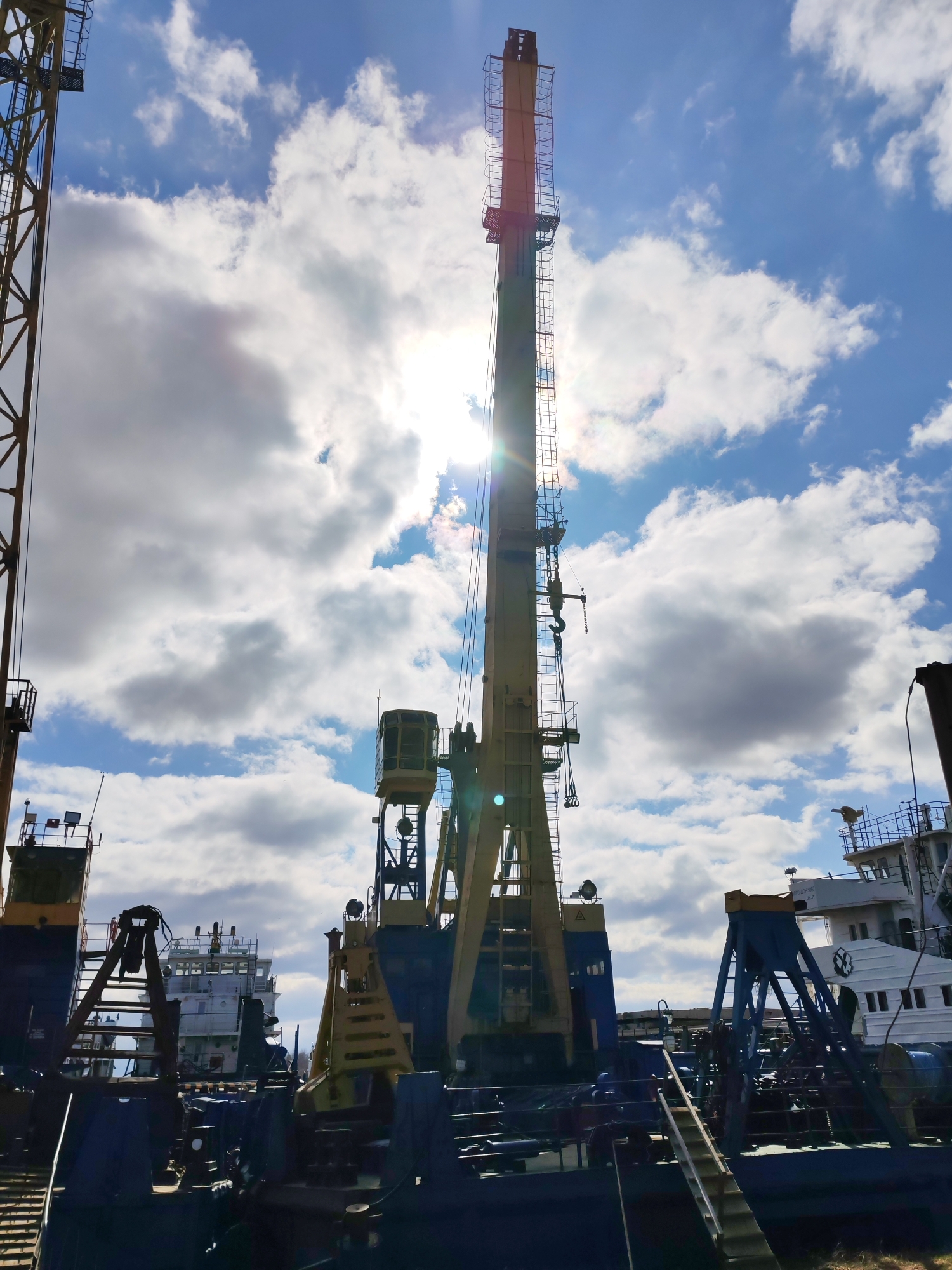 The image shows a view of a harbor area in Rybinsk, Russia, with various cranes and boats. The main subject is a large crane with a yellow and blue color scheme, its extended arm reaching high into the sky, partially obscuring the sun. Other cranes are visible on the left side of the frame. In the background, there are several boats docked, including a white vessel with visible windows and details. The sky is blue with puffy white clouds.