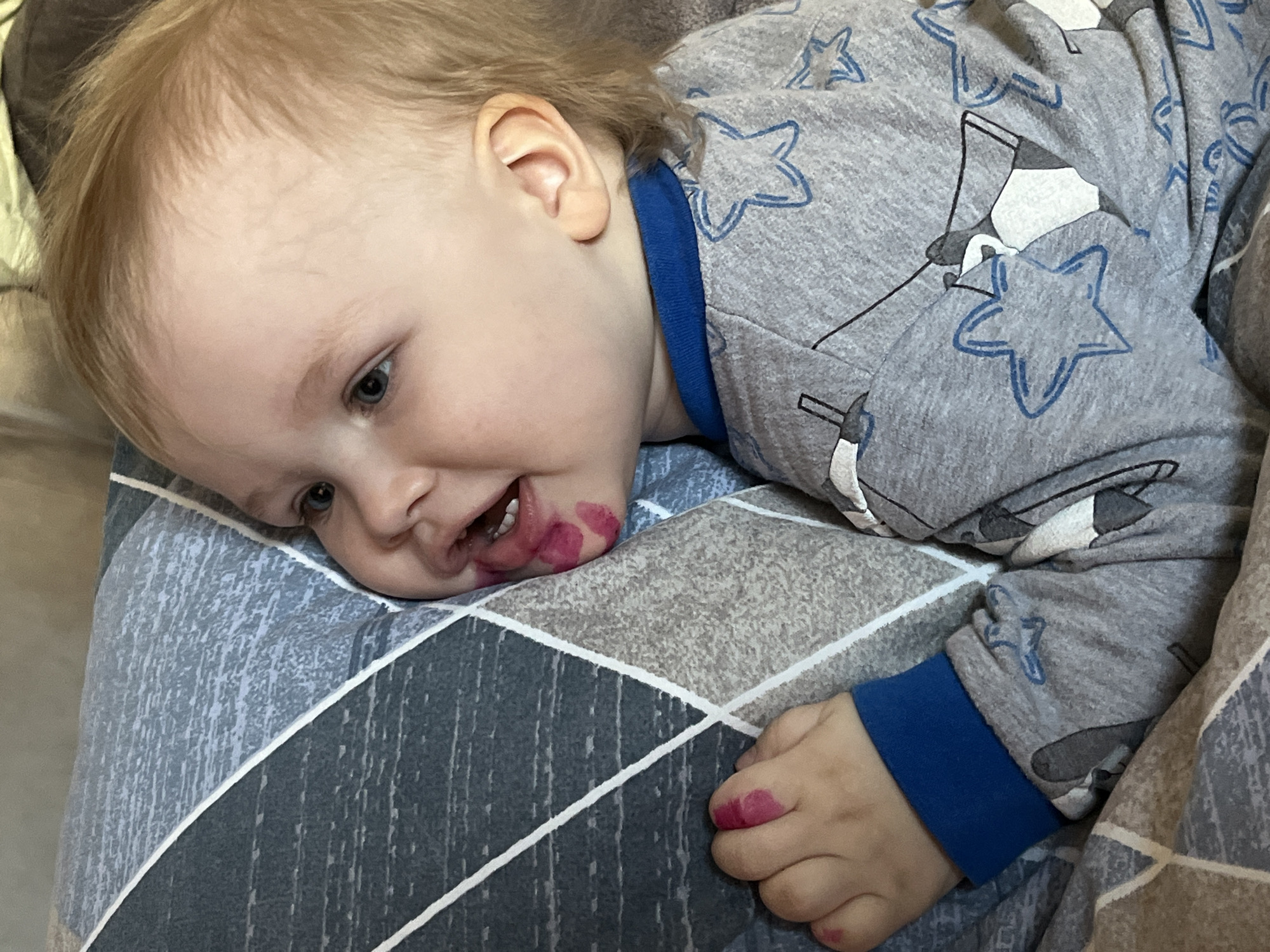 A toddler with blonde hair is lying on a blue and gray patterned fabric, possibly a blanket or pillow. The child has pinkish-red stains around their mouth and on their hand, suggesting they may have gotten into something like lipstick or a marker. The child is wearing a gray pajama top with blue trim, featuring blue stars and possibly penguin-like designs. The exact location in the city of Chaikovsky, Russia cannot be determined from the image.