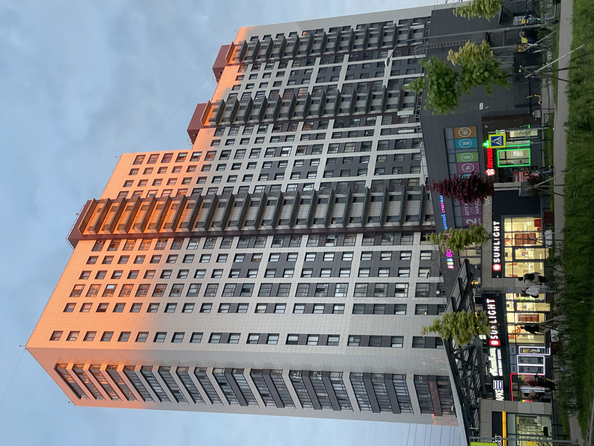 The image displays a tall, modern residential building with commercial establishments on its ground floor, set against an overcast sky during what appears to be late afternoon or early evening. One side of the building is illuminated by the warm, orange glow of the setting sun, creating a strong contrast with the cooler tones of the sky and the shaded portions of the structure.
The building exhibits a contemporary design with a facade composed of varying shades of grey and white panels, featuring numerous windows and recessed balconies. Some sections of the building appear to be stepped or terraced.
At the street level, a row of shops is visible, with interior lights already switched on. Several individuals can be seen near these storefronts, suggesting ongoing commercial activity. Visible text on the shop signs is predominantly in Cyrillic. Prominent signs include a yellow one for "ПЯТЁРОЧКА" (Pyaterochka, a supermarket chain), two green signs for "SUNLIGHT" (a jewelry store chain), and signs for "АПТЕКА" (Pharmacy) and "ДЕТСКИЙ СУПЕРМАР..." (Children's Supermar...). Another sign displays "888 BO...".
Small green trees line the sidewalk in front of the shops, and a street lamp is also present. The scene captures a bustling urban environment characterized by modern architecture and commercial activity.