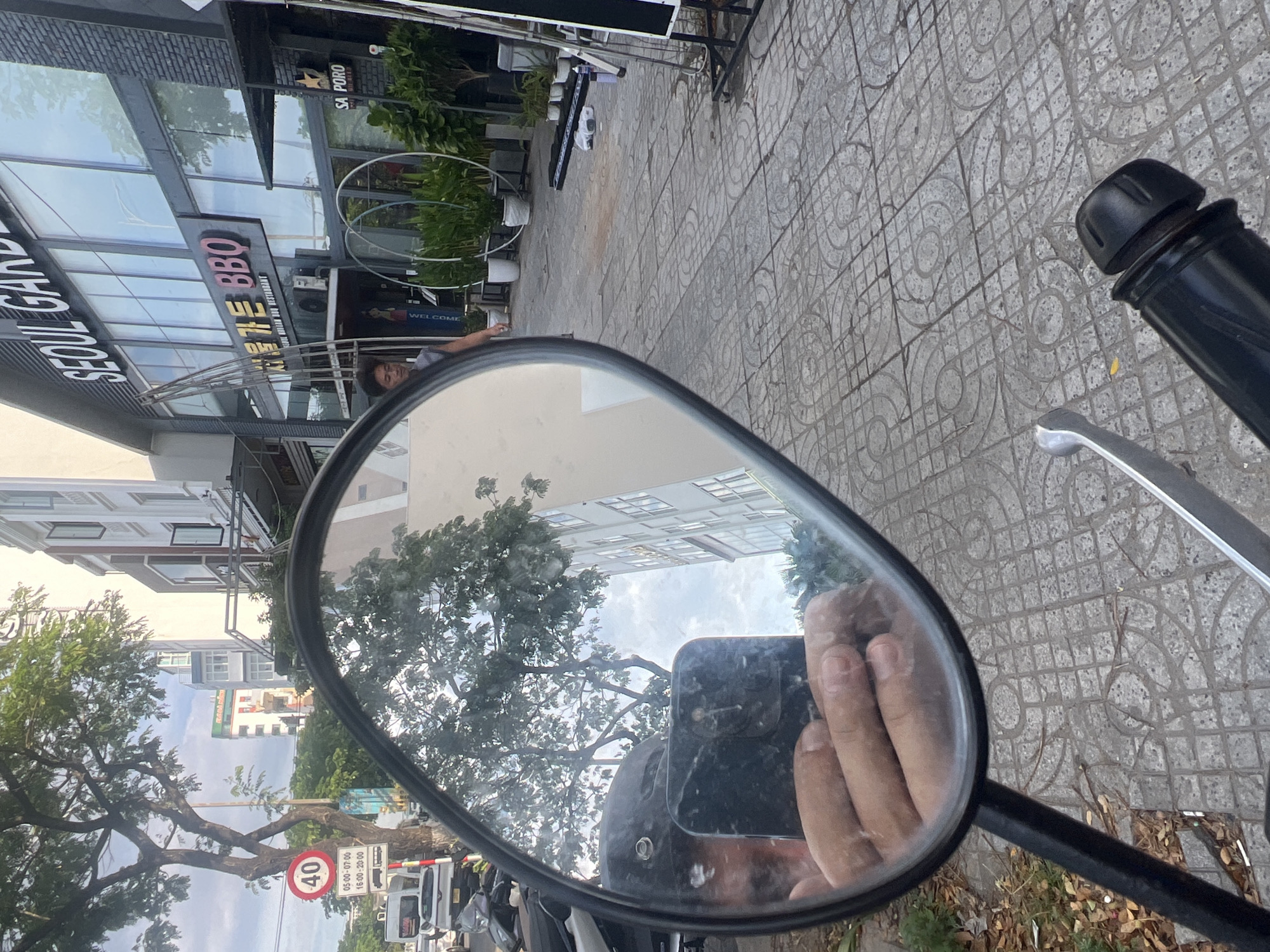 This is a first-person perspective photograph taken from a motorcycle in the city of Đà Nẵng, Vietnam. The rider's hand and phone are reflected in the motorcycle's rearview mirror. In the background, there is a sidewalk with a distinctive tile pattern, a street, and buildings. One building prominently features the signage "SEOUL GARDEN BBQ" in both Korean and English, indicating a Korean restaurant. Another building has a sign that appears to be "SAPORO". A man is visible walking on the sidewalk near the restaurant entrance. A traffic sign indicating a speed limit of 40 is also visible on the left side of the image. The scene appears to be during daylight.