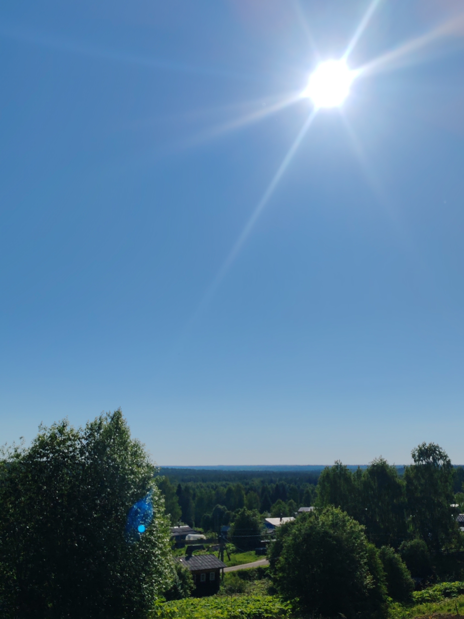 The image captures a sun-drenched rural landscape under a clear blue sky in Чухлэм, Russia. The sky, occupying the upper two-thirds of the frame, is a uniform light blue. A brilliant white sun, emitting prominent starburst rays, is positioned in the upper right quadrant. A distinct circular blue lens flare is visible in the lower-left quadrant, superimposed over the dense green foliage of a large tree that occupies the lower-left foreground. In the midground, several scattered green trees and dark-roofed structures, consistent with village houses or sheds, are discernible. A light-colored ground path or road winds through this area. The background features an extensive, dark green forest stretching across the horizon, meeting a lighter, hazy blue sky in the distance. No human figures or specific actions are visible.