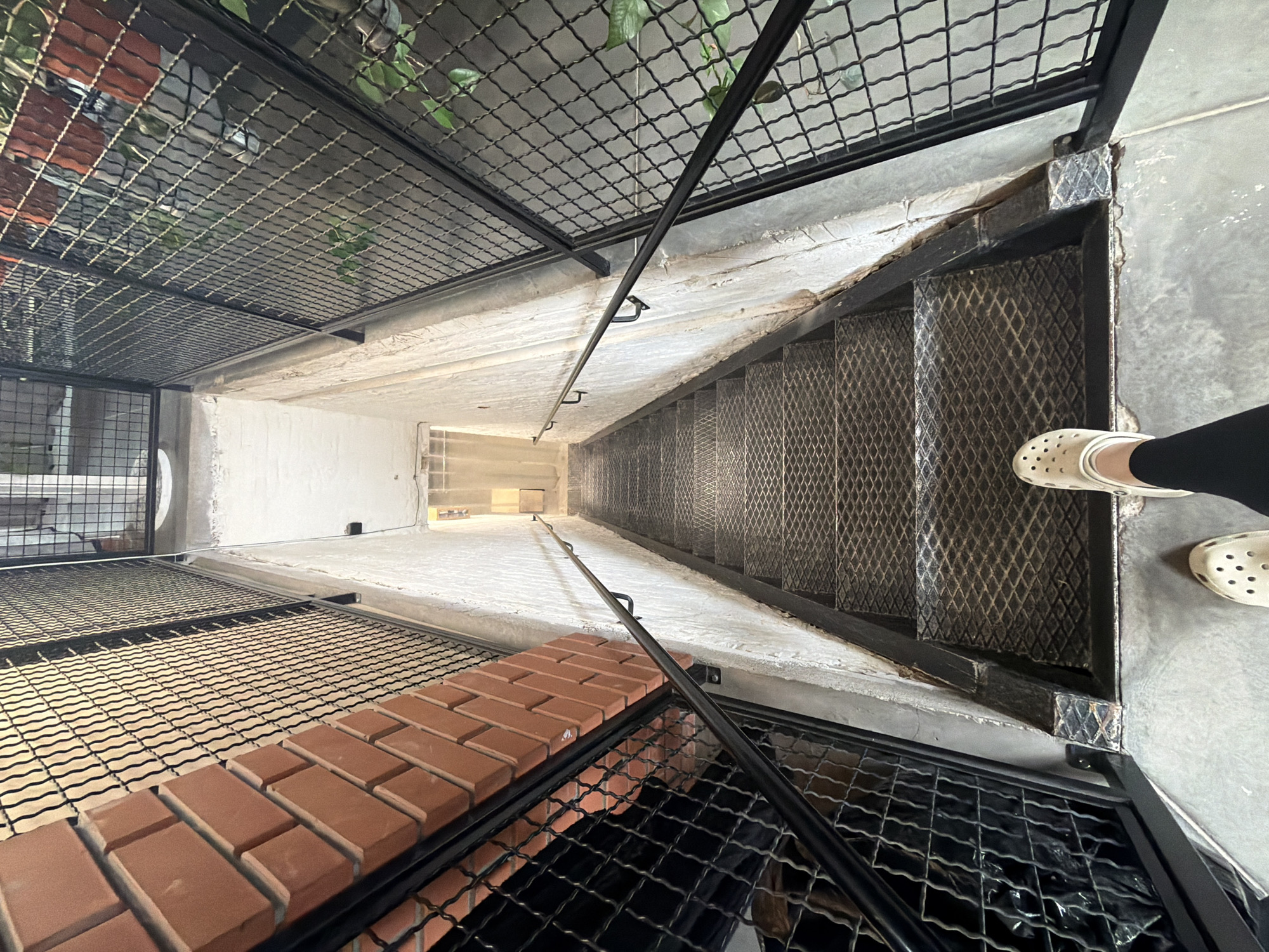 An interior view looks down a set of industrial-style metal stairs. The stairs are constructed with diamond plate treads and risers. A black metal handrail runs along the right side of the staircase, attached to a rough, textured concrete wall. The left side of the staircase is bordered by a black metal mesh fence, behind which is a wall of orange bricks. The perspective is from the top of the stairs, looking down, with a person's legs and feet, wearing white Crocs and black leggings, visible at the bottom of the frame. Green plants in terracotta pots are visible through the mesh fence on the right side of the stairs. The environment suggests an urban, possibly commercial or industrial, setting.