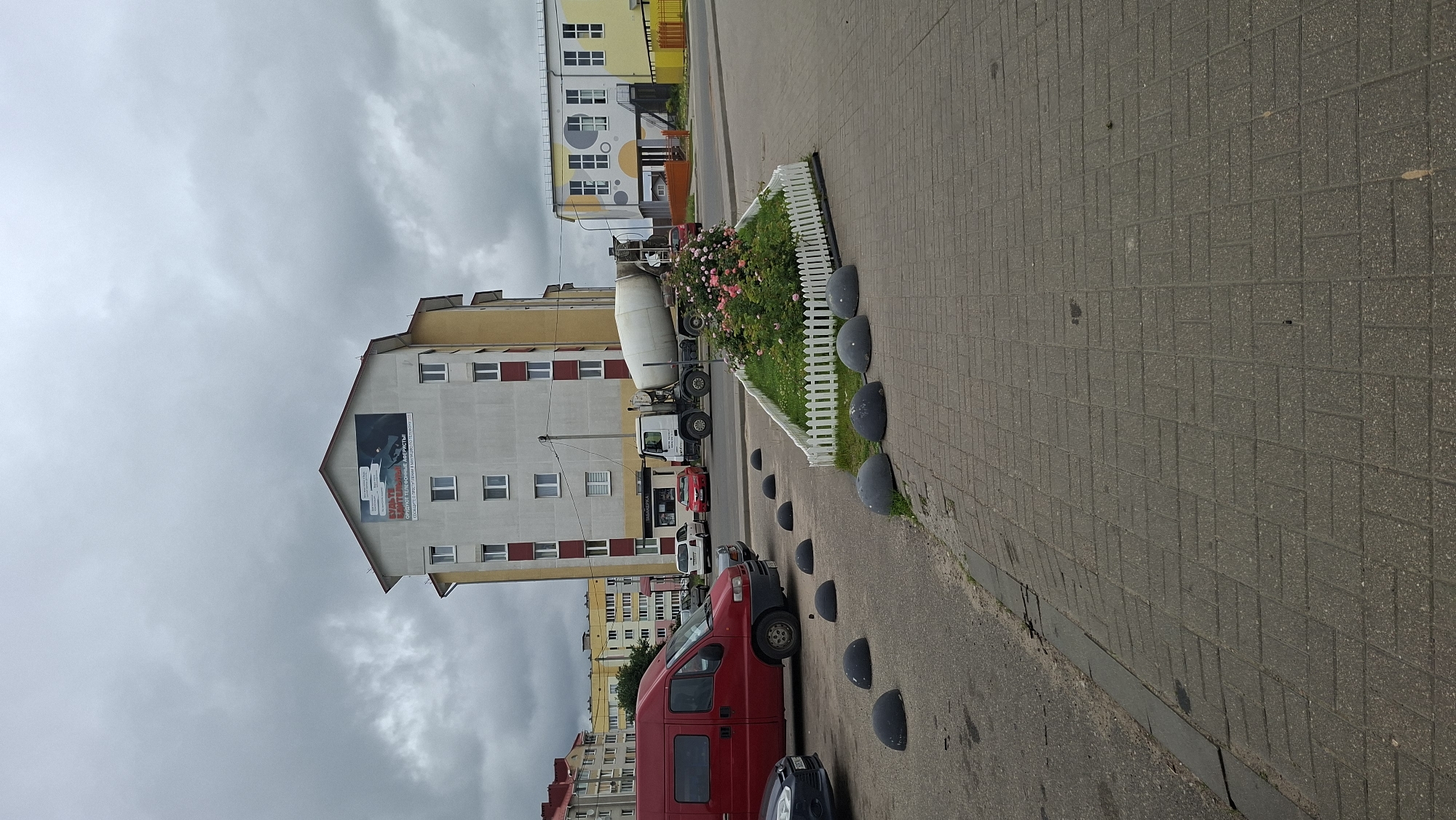 In the city of Smalyavichy, Belarus, on a cloudy day, a street scene unfolds. A red commercial van is parked on the left, facing away from the viewer. To its right, a white cement mixer truck is positioned, its drum angled towards the right. In front of the truck and partially obscured by it, a triangular flower bed with pink roses and green grass is enclosed by a low white picket fence. Several dark, rounded bollards are placed in a line along the edge of a paved pedestrian area in the foreground.
Behind the flower bed and the truck, a multi-story building with a light-colored facade and yellow accents dominates the background. A large advertisement is displayed on the upper section of this building. Further to the right, another building with a yellow and white facade, featuring circular window designs, is visible. The sky is overcast with thick, gray clouds. The environment appears to be an urban setting with pavement and buildings characteristic of a city.