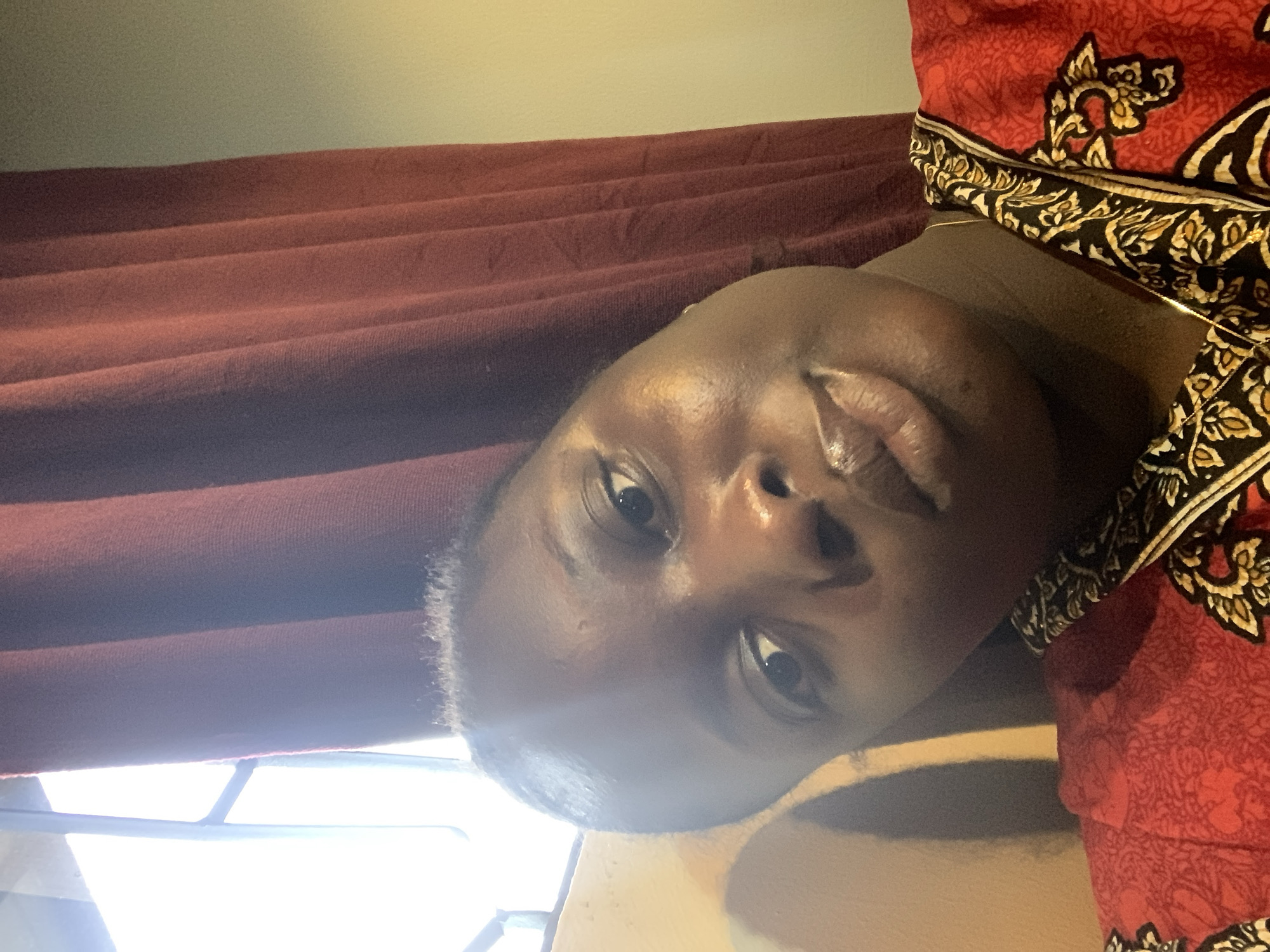 A close-up, head-and-shoulders selfie captures a young Black woman tilted to her right, with her face angled towards the viewer. Her eyes are open, looking slightly to her left, and her lips are slightly parted. She wears a vibrant red garment adorned with a black, white, and gold patterned trim around the neckline. The background consists of a deep red curtain on the right and a bright, out-of-focus window on the left, suggesting an indoor setting with natural light. The image is likely taken in an urban environment.