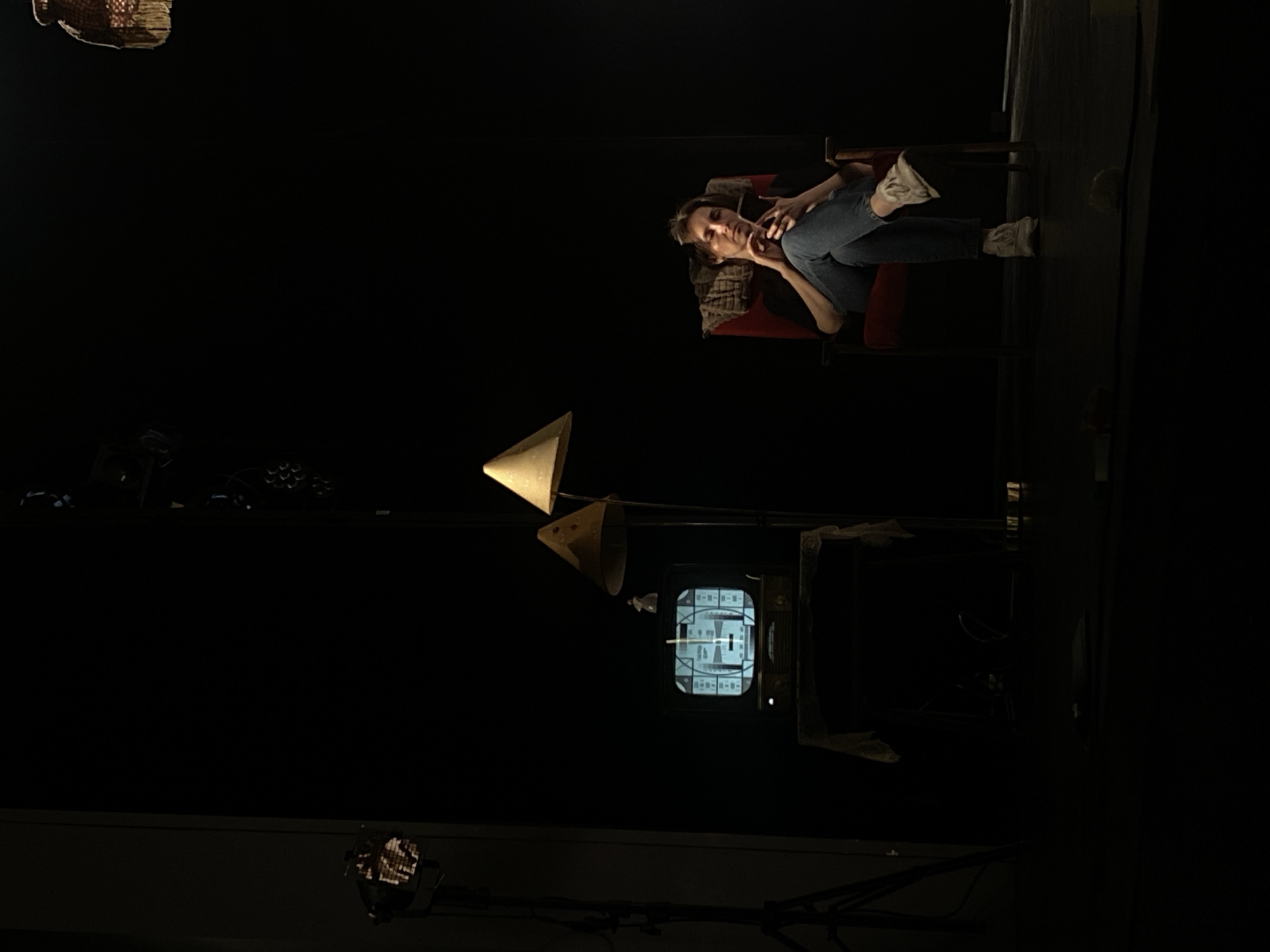 The image shows a dark room. A woman lies reclined in a red chair, wearing jeans and sneakers. An old television set displaying a test pattern sits on a stand with a cone-shaped lamp positioned nearby. Other objects, including a stage light, are partially visible. It is impossible to identify the location as the city of Zelenograd, Russia from the context of this image.