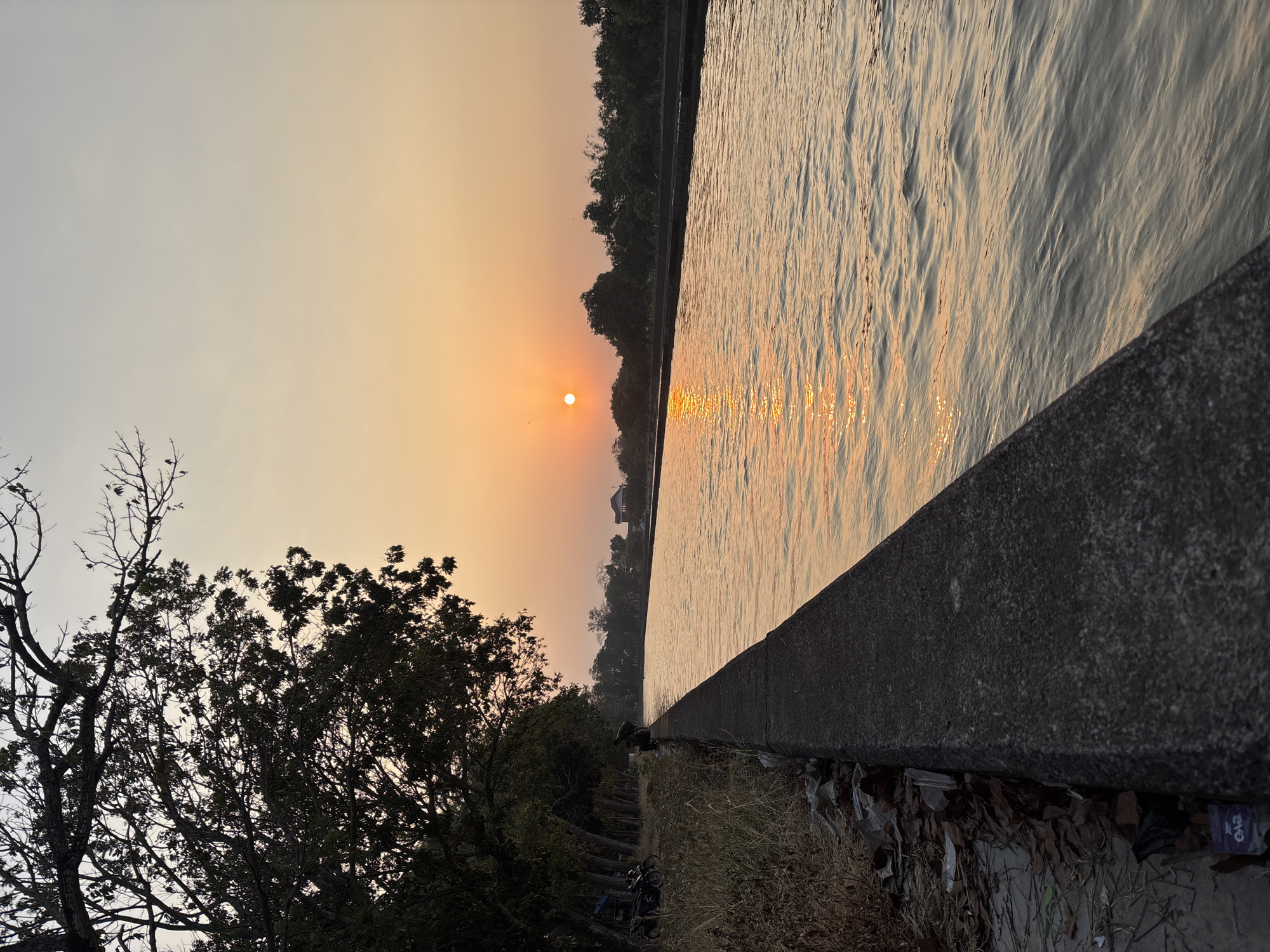 The image depicts a serene canal scene at sunset in Panyingkiran, Indonesia. The sun, a bright orange orb, is low in the sky, casting a warm glow that reflects on the rippling water. Tall trees line the left bank, their silhouettes stark against the fading light. A concrete embankment runs along the water's edge, with dry grass and debris scattered on the bank. A few people are visible near the embankment, one sitting and two others near motorcycles. The right bank is also lined with trees and in the distance, a building with a distinctive roof can be seen. The overall atmosphere is peaceful, marked by the soft hues of dusk.