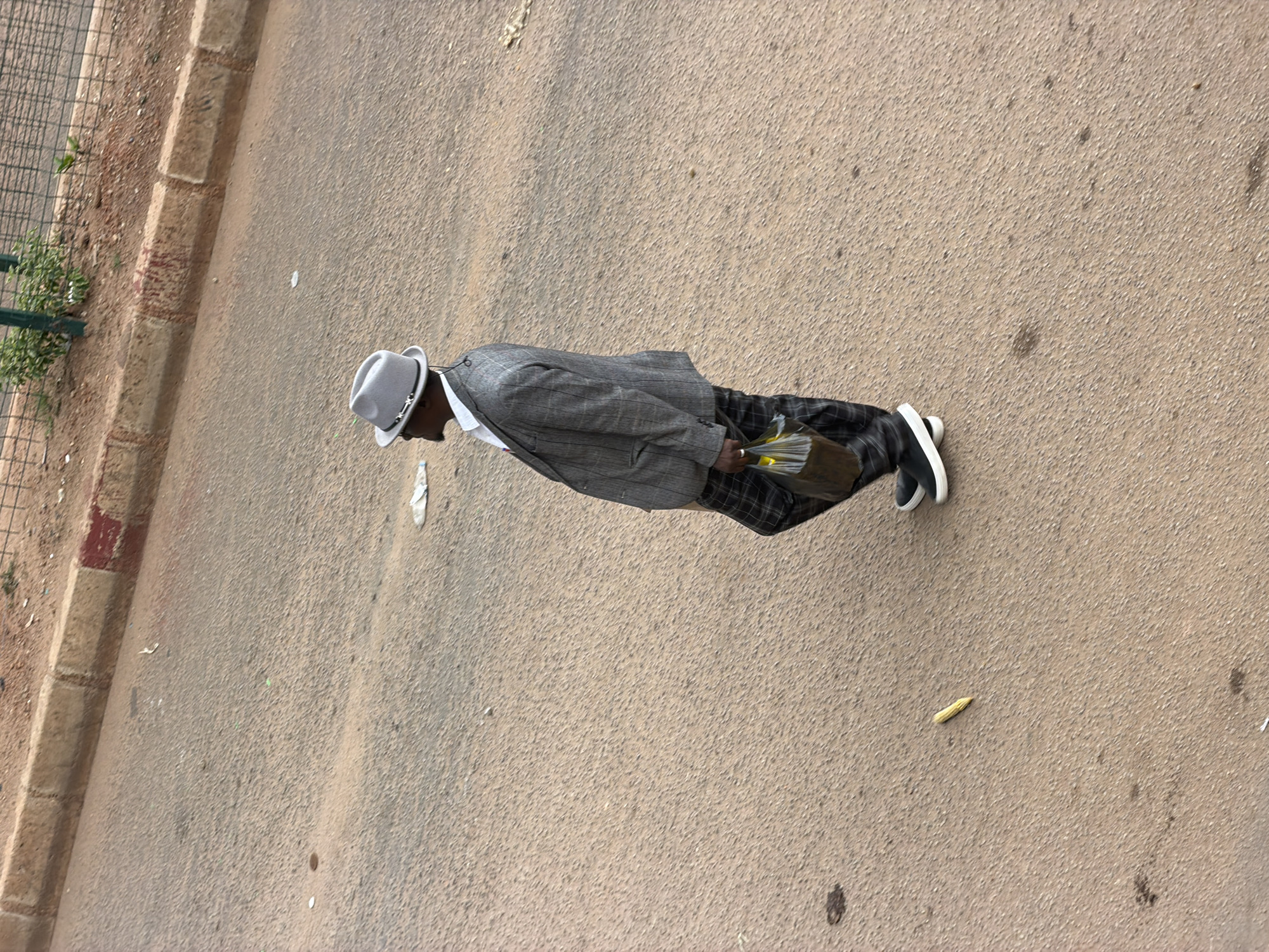 A lone Black male wearing a light grey fedora hat, a pinstripe suit jacket, a white dress shirt, and plaid trousers walks on a paved road in Bukuru, Nigeria. He is carrying a clear plastic bag containing some items in his right hand. A single piece of corn is visible on the road in front of him. The road appears to be asphalt with some reddish-brown dirt or sand. A curb with red and white markings is visible on the left side of the image, and a metal fence with some greenery behind it is in the background.