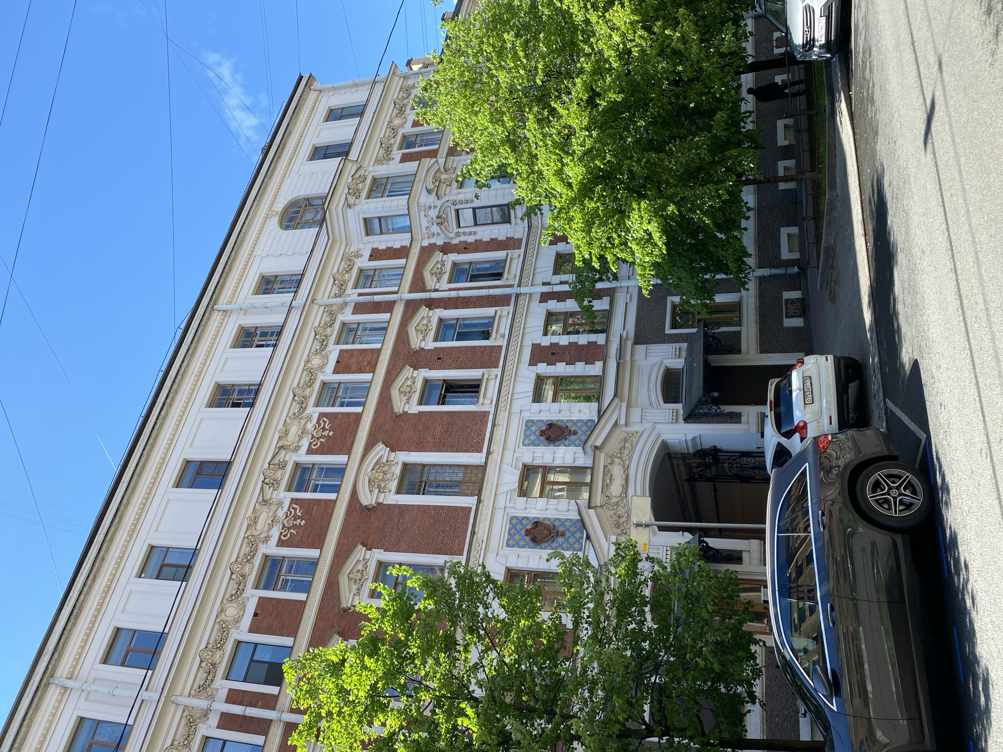 The image shows a multi-story building with ornate architectural details, including red brick accents, white trim, and decorative window frames. The building has multiple windows on each floor. Lush green trees are visible in front of the building, partially obscuring the lower levels. Two cars are parked on the street in front of the building, one white and one dark-colored. A narrow street is visible to the right of the cars. Overhead power lines stretch across the upper portion of the image against a clear blue sky. The building's architecture and street scene suggest an urban environment.