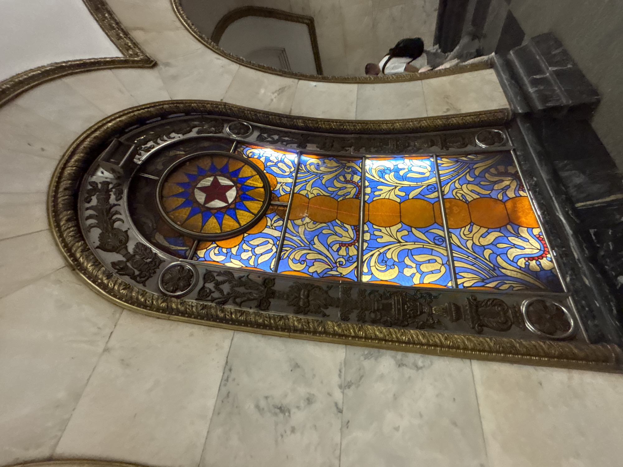 The image depicts a stained-glass window in what appears to be a public building, possibly in Moscow, Russia, given the presence of a red star motif. The window is ornate, featuring a central circular design with a red star in the middle surrounded by blue and yellow glass. The rest of the window consists of panels with blue backgrounds and swirling yellow and gold floral patterns. The window is set in an elaborate dark metal frame with decorative elements.
The window is situated in a wall covered in white marble tiles. A portion of a second-floor balcony can be seen above, where two people are visible. The scene seems to be inside a building with high ceilings and classic architectural details.