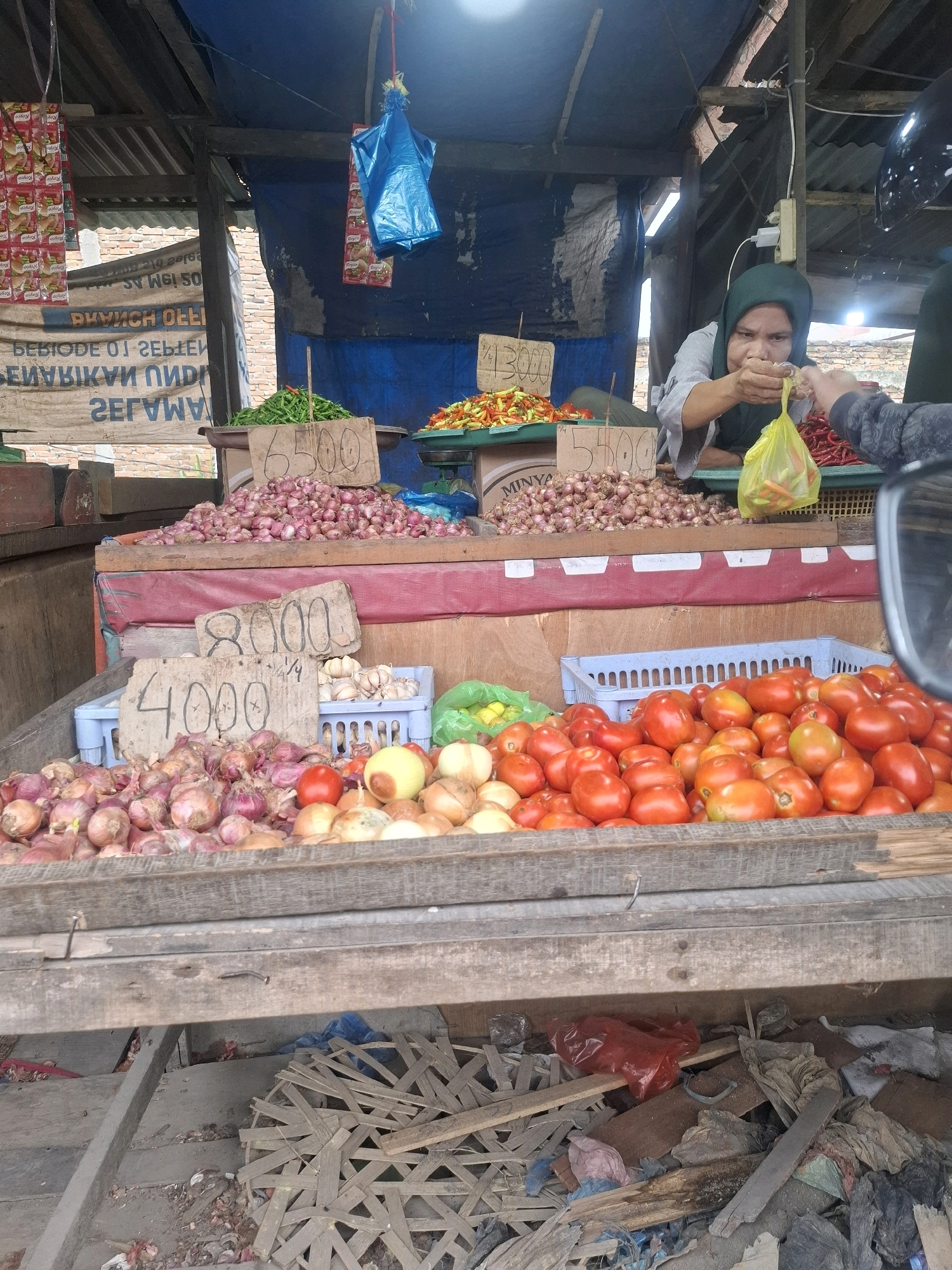 A street market stall in Kenangan, Indonesia, features a woman in a green headscarf weighing chili peppers in a yellow plastic bag for a customer. The stall displays a variety of fresh produce, including heaps of red onions, garlic, tomatoes, and various types of chili peppers. Wooden crates and signs with handwritten prices are visible. The background shows a rustic, open-air market structure with tarpaulin roofing and brick walls. The ground is littered with debris, including discarded wood and plastic.