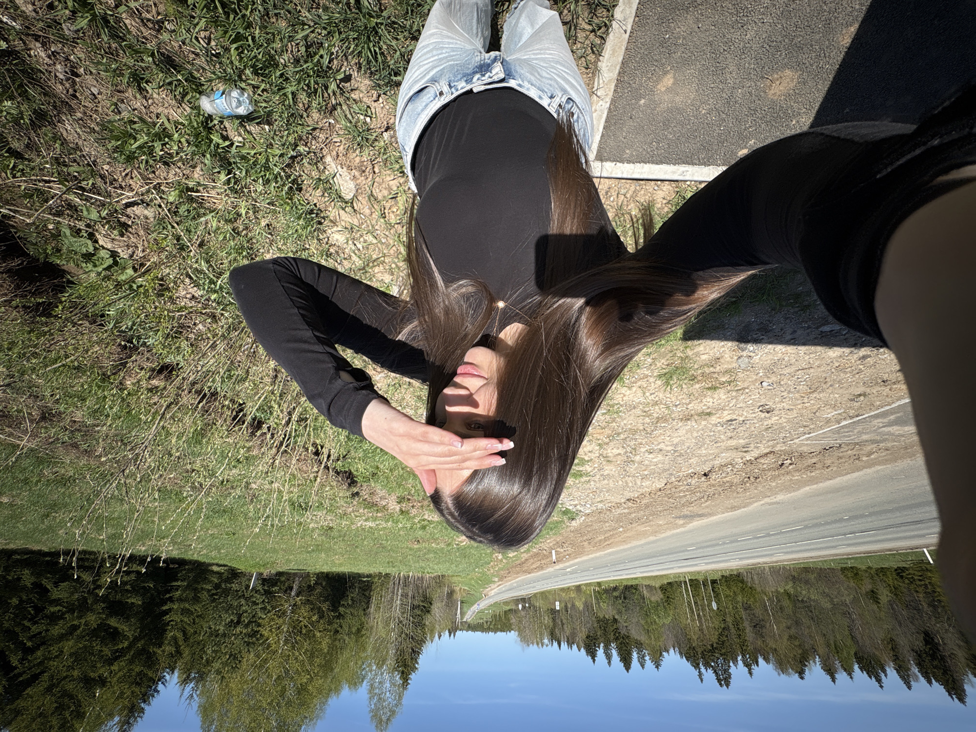 A woman stands on the side of a road in Stepanovka, Russia, obscuring her face with her hand. She wears a black long-sleeved shirt and light-wash jeans. A discarded plastic bottle lies on the grassy shoulder. The road stretches out in front of her, lined by a forest that reflects onto the clear blue sky.