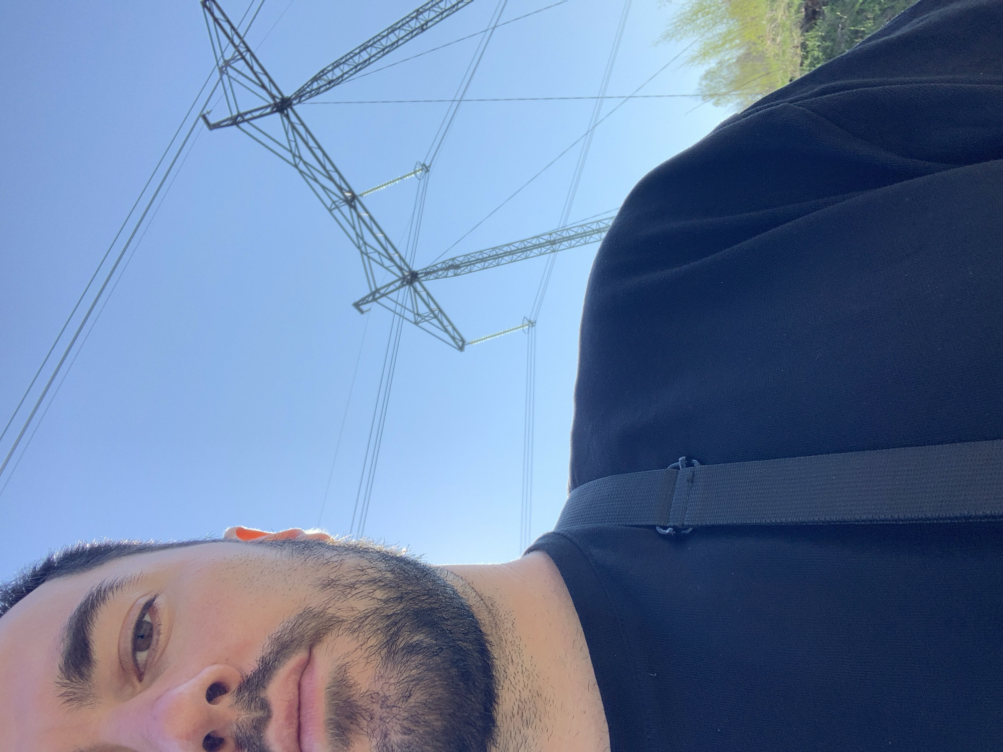 The image is a selfie taken outdoors in Chaikovsky, Russia. A man with dark hair and a beard is partially visible in the foreground, his face tilted. He is wearing a dark-colored shirt. Behind him and above is a clear, blue sky with high-voltage power lines and transmission towers. The environment appears to be an open area with some vegetation visible in the distant background. A strap is seen across the man’s shirt.