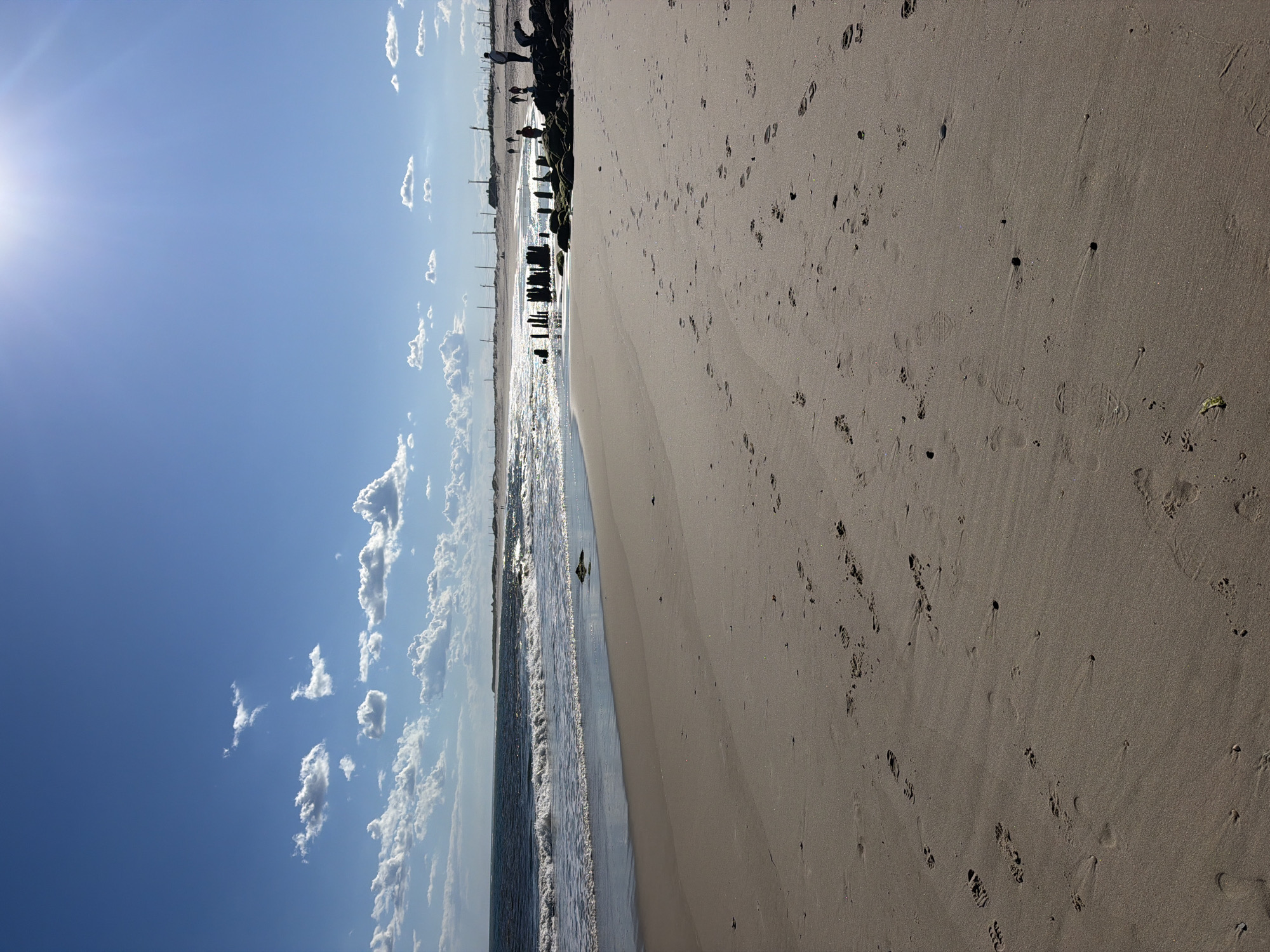 An eye-level, vertical, wide shot captures a sunlit beach scene on a clear day, with the sun's glare in the upper right corner. The foreground is dominated by sand marked with numerous footprints, indicating recent human and possibly animal activity. The midground features the ocean with gentle waves lapping at the shore. Along the water's edge, a few rocks and weathered wooden pilings stand as remnants of a past structure. In the background, a sandy dune line is visible, topped with a few scattered structures and utility poles, suggesting the proximity of a developed area. Several figures, appearing as small silhouettes, are scattered along the shoreline in the distance, engaged in activities like walking and standing. The sky is a vibrant blue, dotted with fluffy white clouds, enhancing the bright and airy atmosphere of the location in Hampton Bays, USA.
