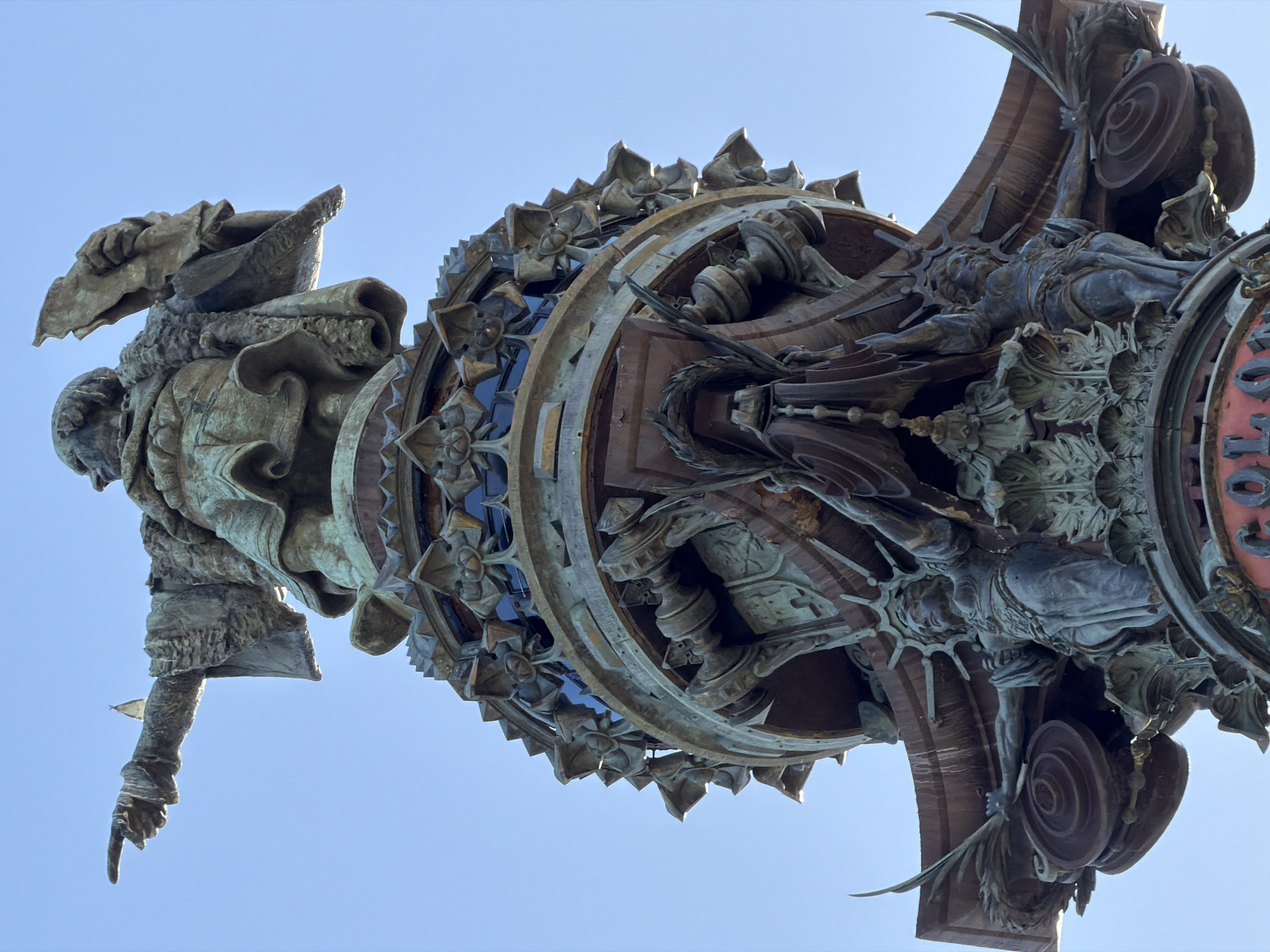 This is a low-angle, close-up, and vertically oriented outdoor photograph of a bronze monument under a clear blue sky. The monument is intricately detailed, featuring multiple figures and ornate architectural elements.
The primary subject appears to be a large, imposing bronze statue of a person, seen from the chest up, wearing a cloak and a pointed hat. The figure is leaning backward, with its right arm extended downward and pointing with a finger. Below this figure, the monument expands into a circular, multi-layered structure adorned with smaller figures and decorative motifs. These include what appear to be armored figures, winged beings, and floral patterns. The metalwork has a patina, suggesting age and exposure to the elements.
The scene is bathed in bright, natural light, indicating it is daytime, likely mid-morning or afternoon, given the intensity of the sunlight. The clear blue sky suggests fair weather. There are no discernible location cues other than the provided context of Barcelona, Spain. The overall impression of the monument is one of grandeur and historical significance.
The visible text on the monument, in red lettering on a circular section, is partially obscured but appears to read "MONU..." followed by other letters, suggesting it might be part of a name or inscription. The orientation of the image and the perspective from below create a dramatic and imposing view of the monument's upper sections.