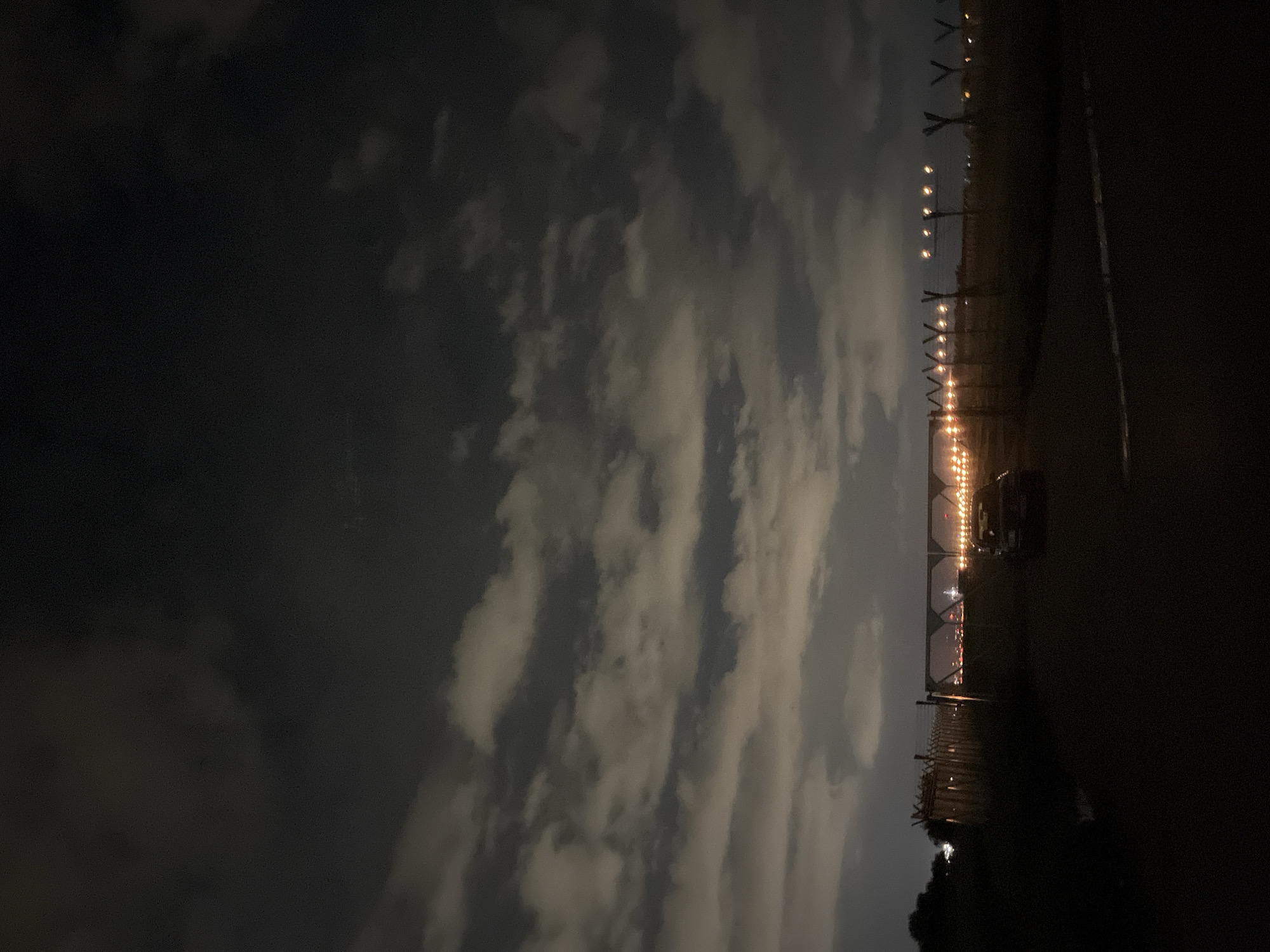 The image shows an outdoor scene at night. On the left, there are clouds illuminated by some light source from below. A line of evenly spaced lights runs horizontally across the middle of the image, disappearing into the distance. To the right of the lights is a dark road. A vehicle is parked near the lights. Based on the details and context provided, the location is near an airport in Sofia, Bulgaria, indicated by the lights along the runway.