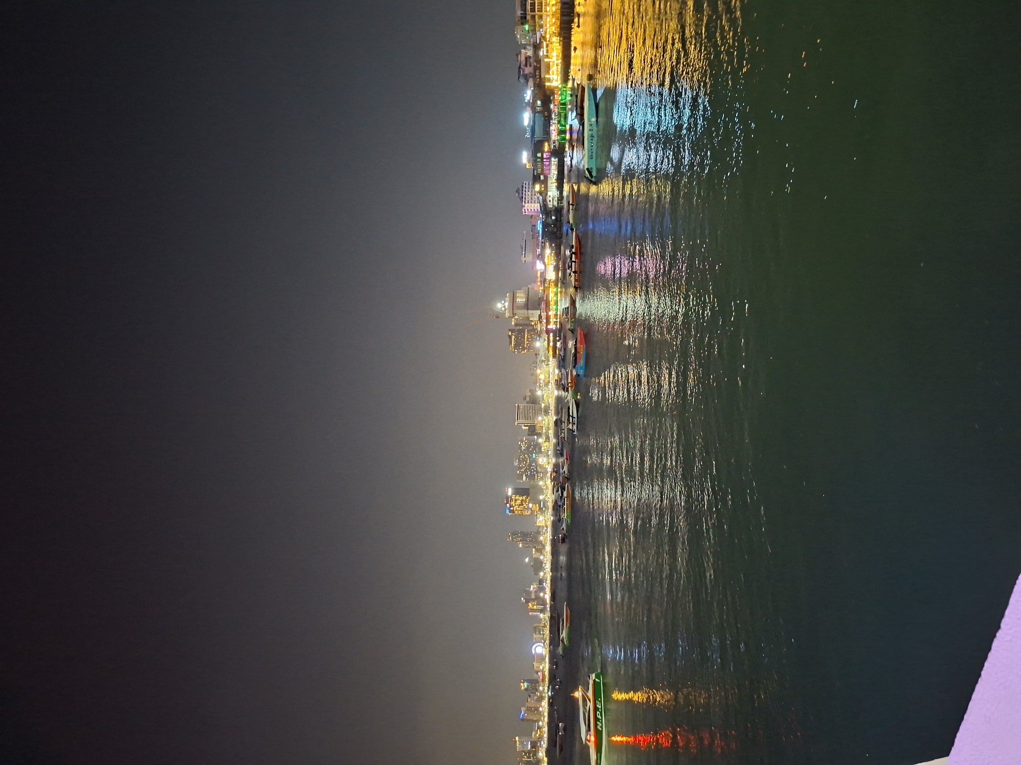 At nighttime in Pattaya City, Thailand, city lights shine brightly, reflecting off the water's surface. Buildings line the waterfront. Boats are visible on the water. The sky is dark and overcast.