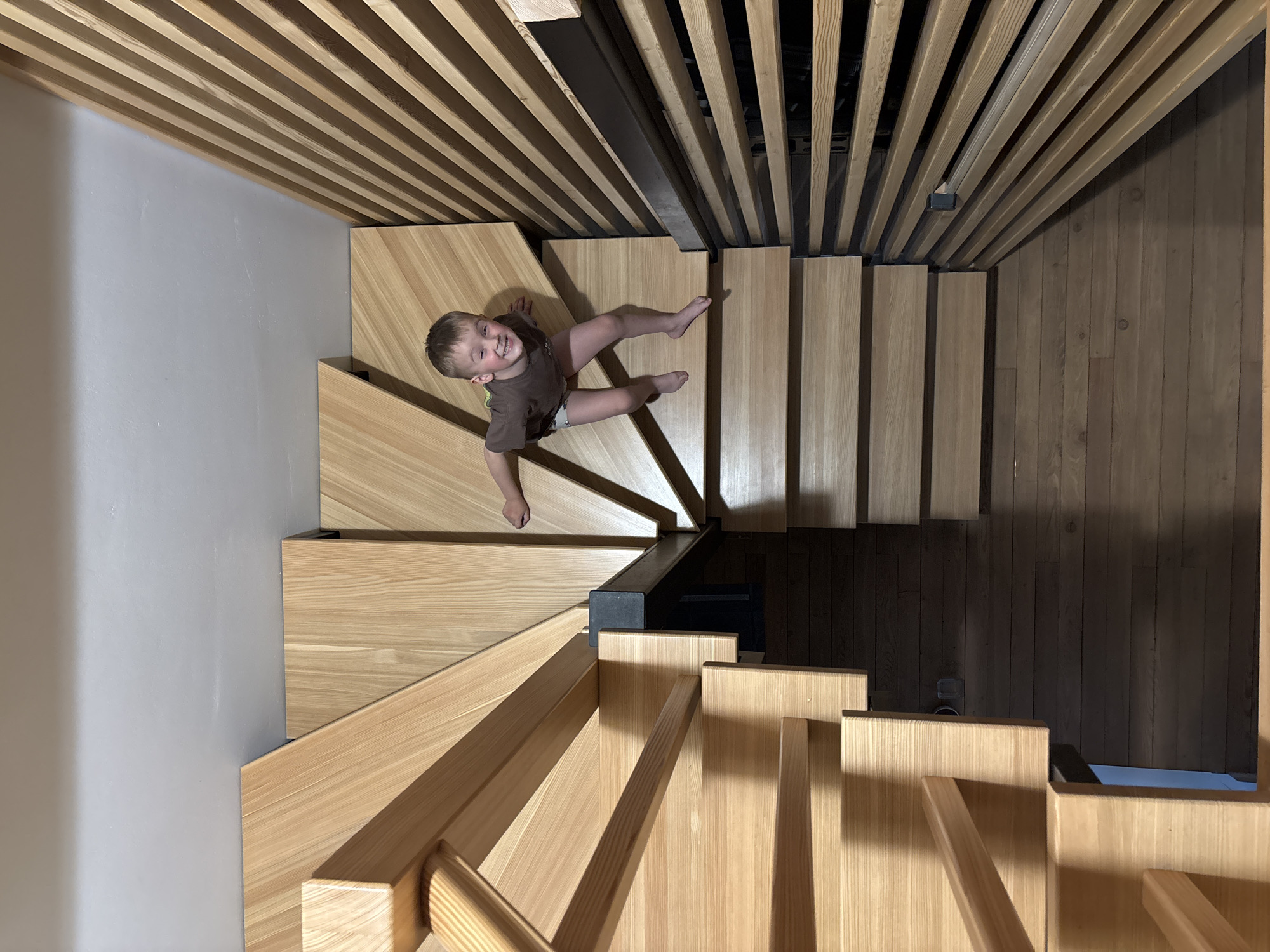 A high-angle shot captures a child seated on a light wood staircase, presumably indoors. The child, wearing a brown shirt and light-colored shorts, is smiling and appears to be looking up at the camera. The staircase has a modern design, with open risers and wooden railings. The ceiling above consists of parallel wooden slats, creating a striped effect. Adjacent to the staircase is a grey wall and a section of darker wood paneling. The location is indicated as Chaikovsky, Russia.